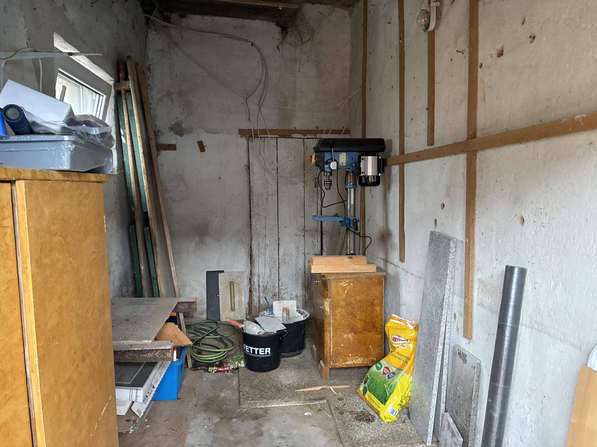 Funktionaler Werkstattraum mit Bohrmaschine, Werkzeugen und Baumaterialien, beleuchtet durch ein Fenster.