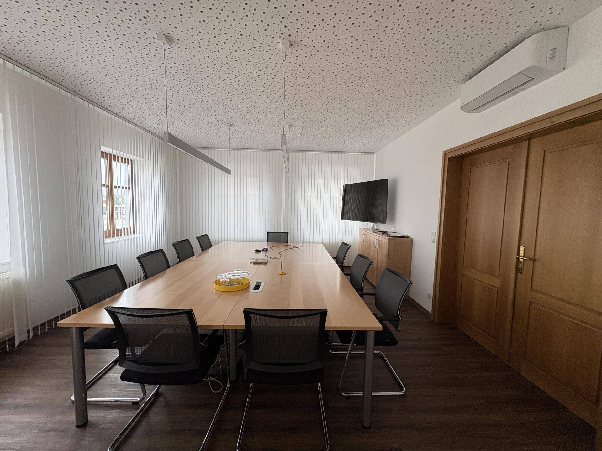 Konferenzraum mit großem Holztisch, vielen Stühlen und einem an der Wand montierten Fernseher.