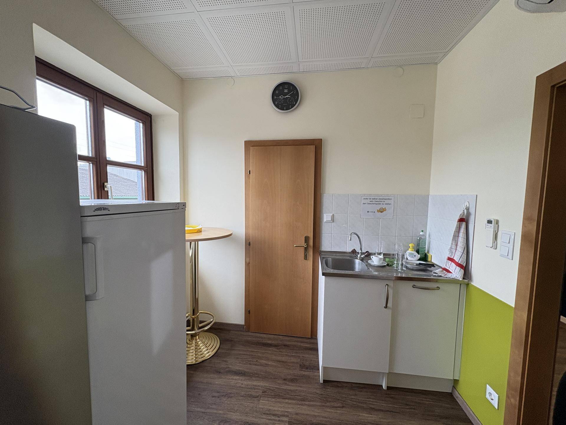 Kleine Küchenzeile mit Spüle, Kühlschrank und einem Stehtisch in einem hellen Raum.