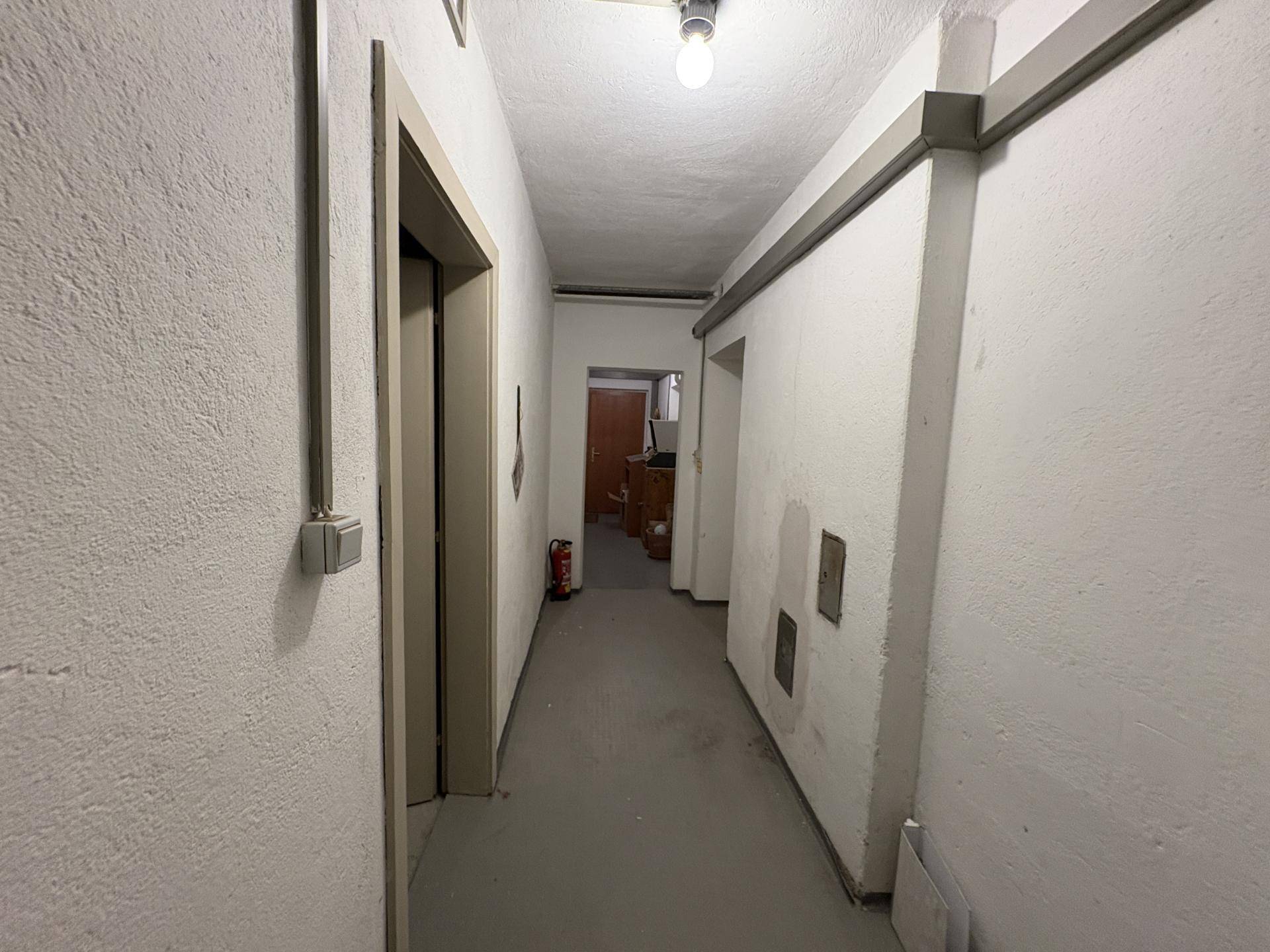 Langer Kellerflur mit einfachen Wänden und Deckenbeleuchtung, führt zu weiteren Räumen.