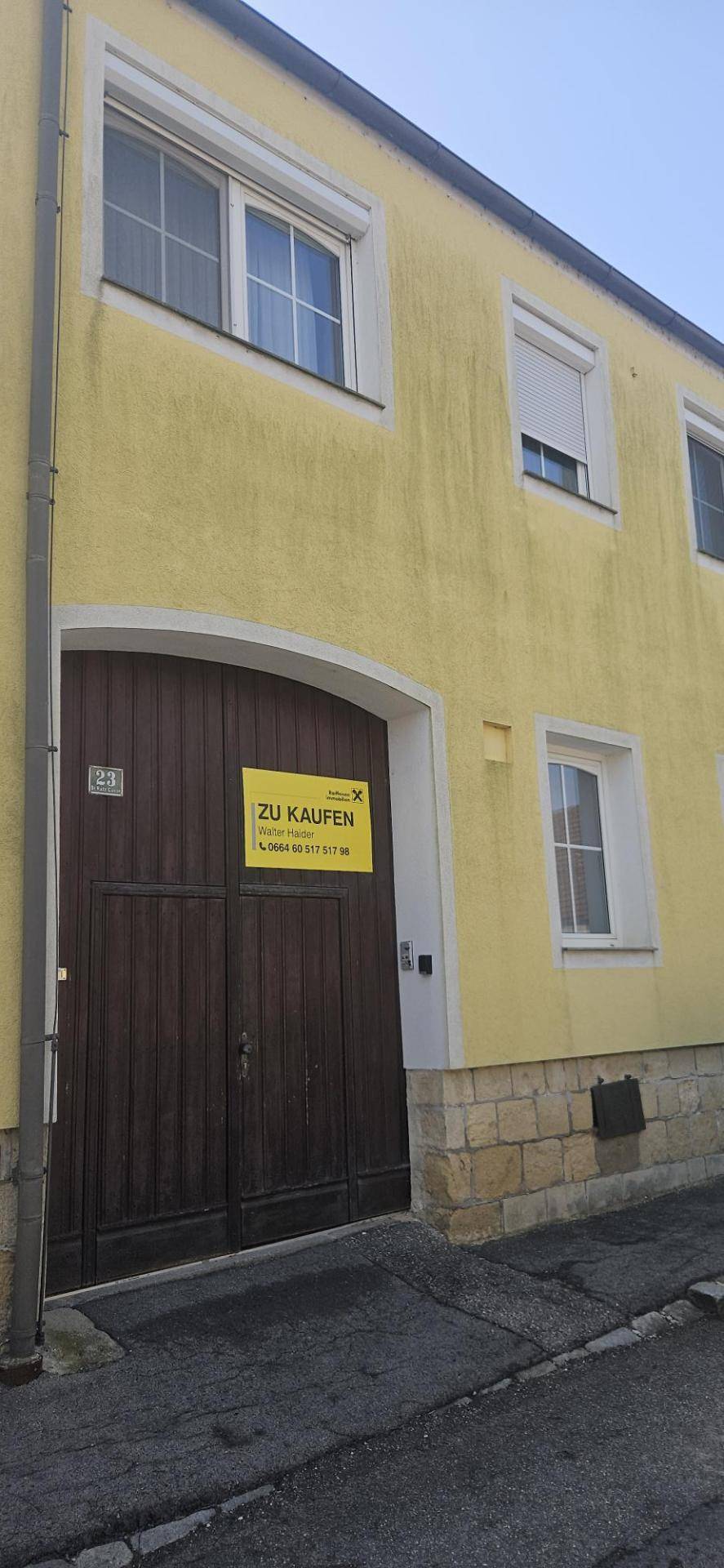 Gelbe Hausfassade mit großem Holztor und Fenstern, ein "Zu Kaufen"-Schild ist angebracht.