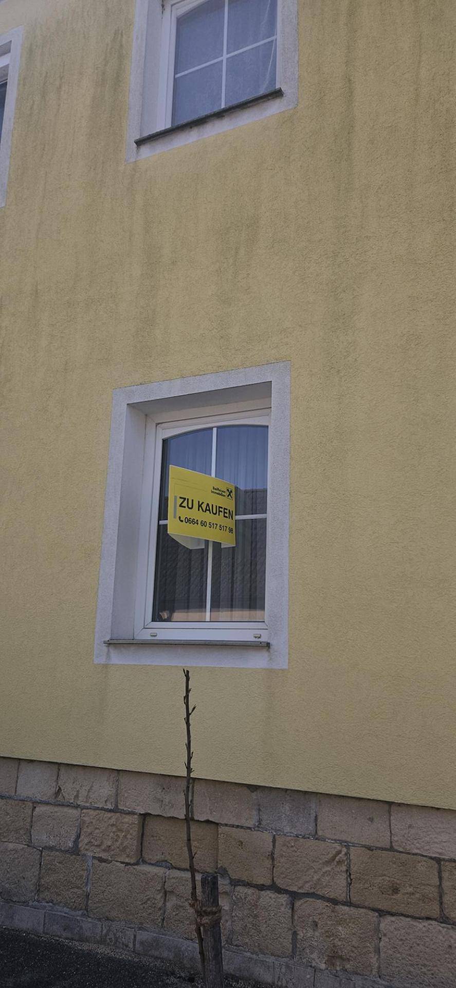 Nahaufnahme der gelben Fassade mit Fenster und einem "Zu Kaufen"-Schild.