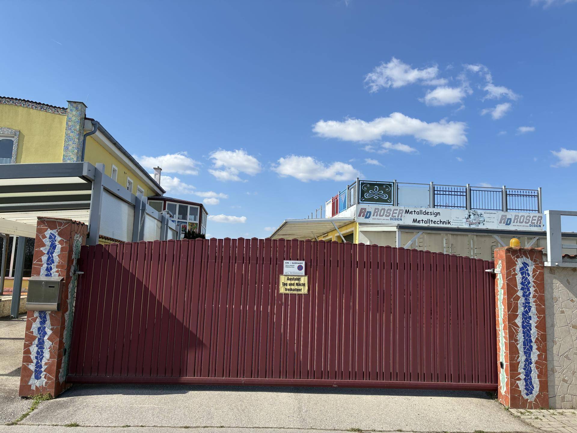 Rotes Eingangstor mit Mosaiksäulen und Blick auf das dahinterliegende Gebäude unter blauem Himmel.