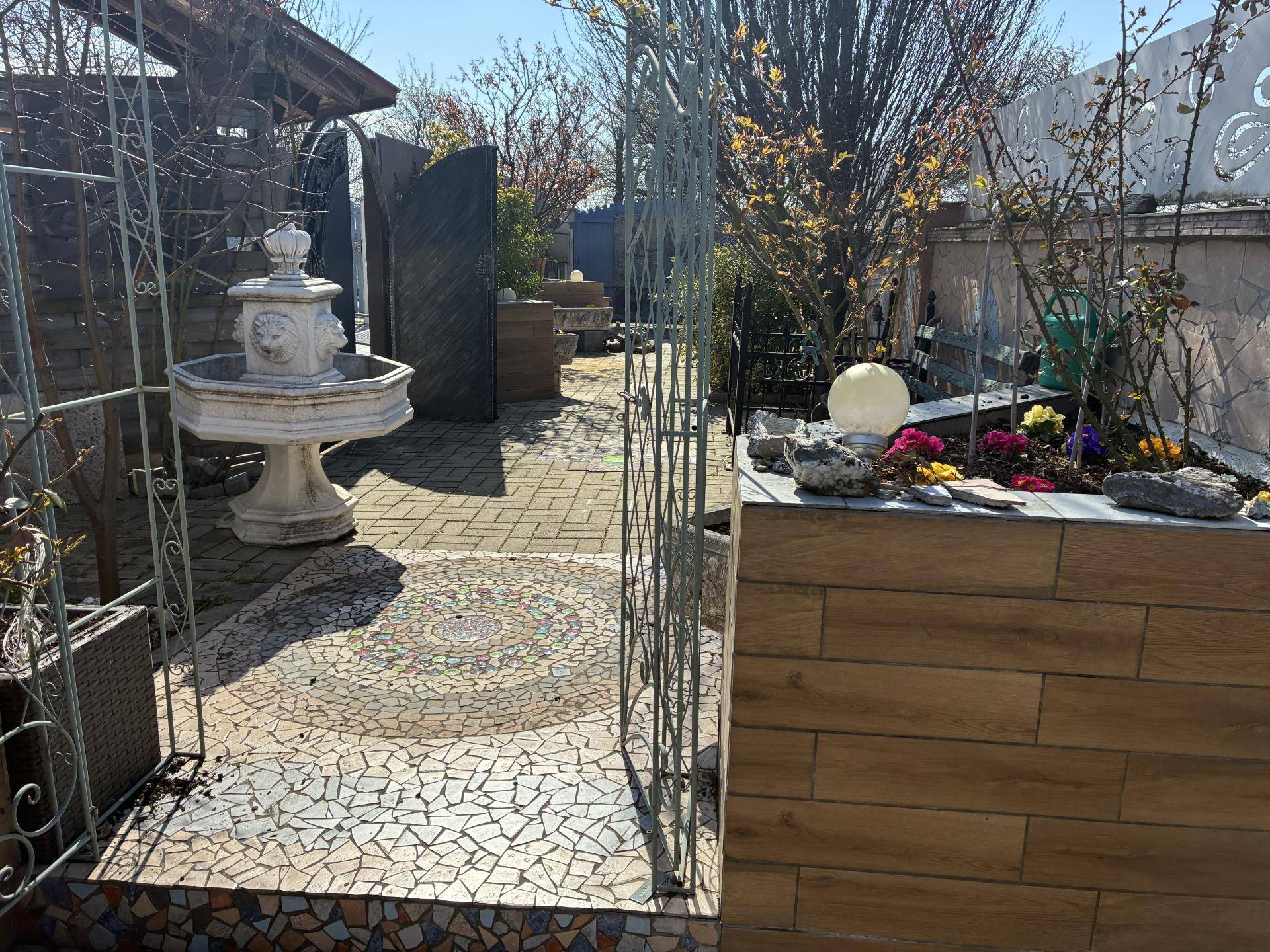 Gepflasterter Gartenbereich mit dekorativem Brunnen, Mosaikfliesen und verschiedenen Pflanzen in Töpfen.
