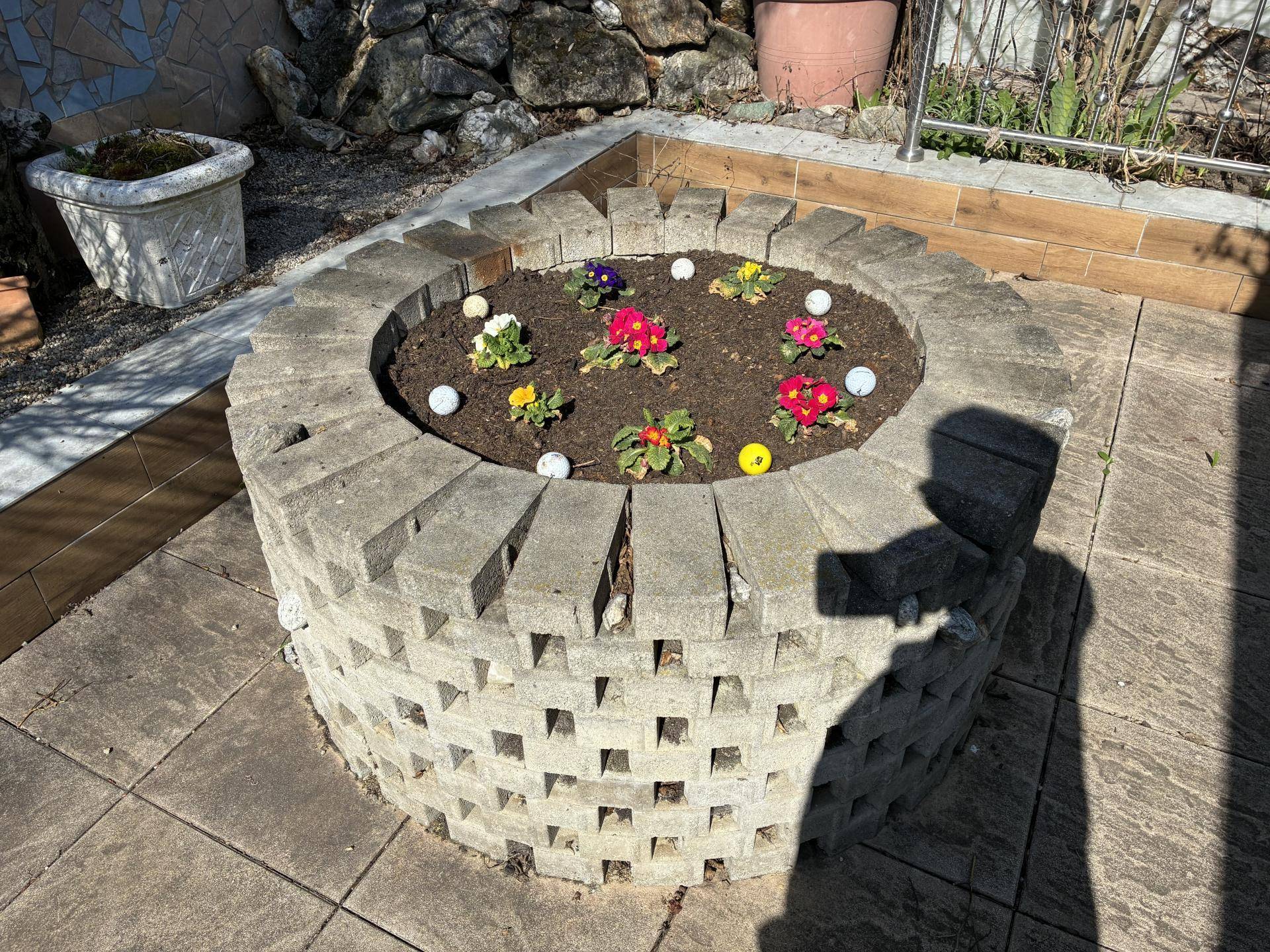 Rundes Hochbeet aus Betonsteinen mit bunten Blumen und kleinen Dekorationselementen auf gepflastertem Boden.