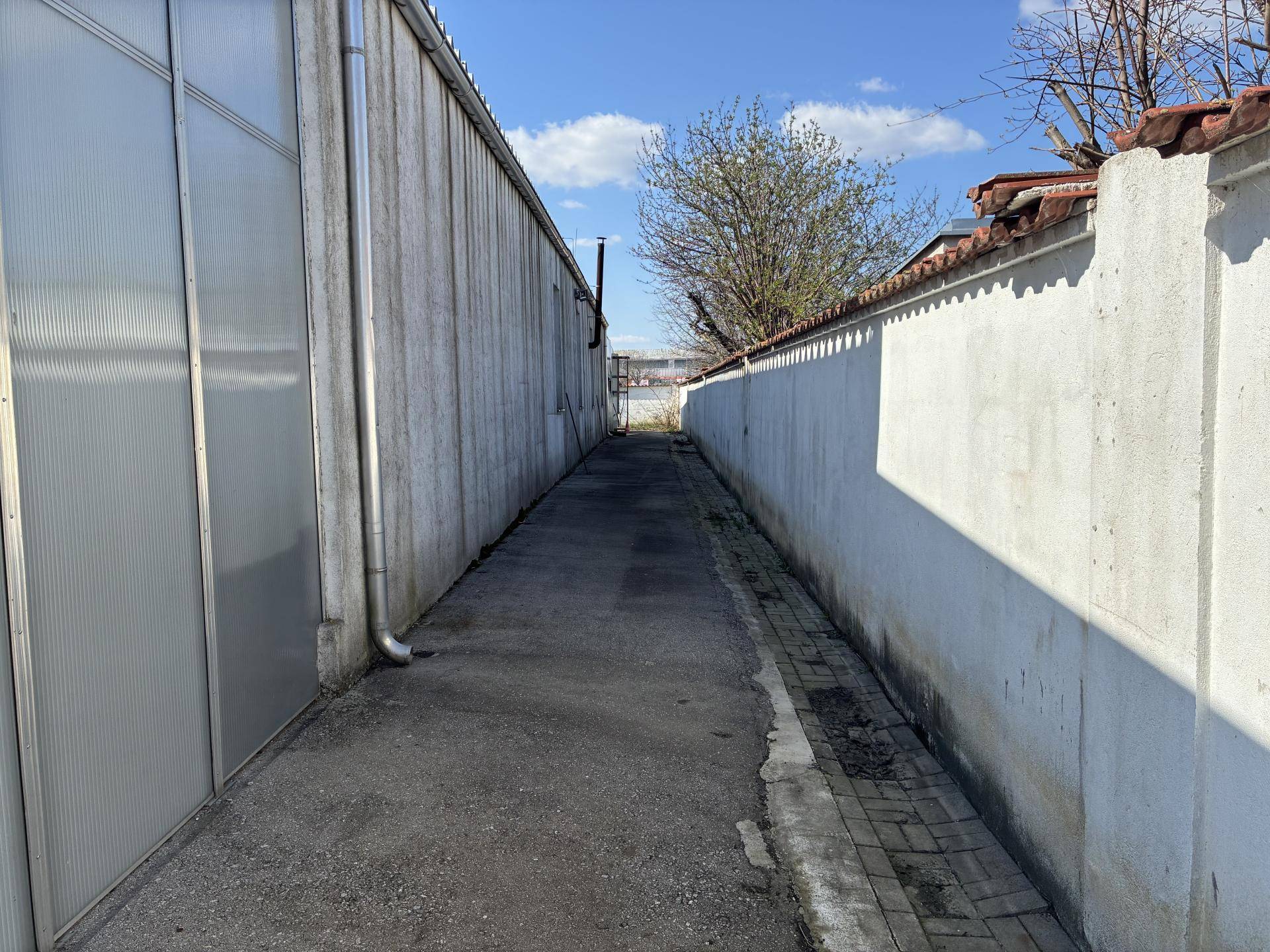 Schmaler Gang oder Weg zwischen zwei Gebäuden, begrenzt von einer weißen Mauer und einer Wellblechfassade unter hellem Himmel.