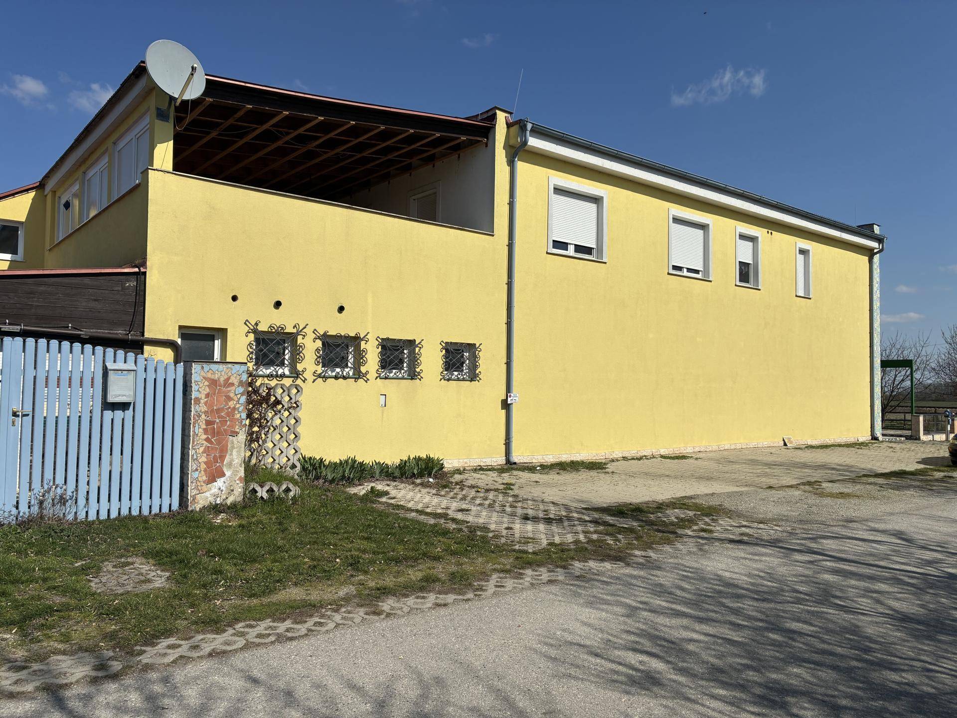 Gelbes Gebäude mit Satellitenschüssel und Rollläden an den Fenstern, davor eine gepflasterte Fläche unter blauem Himmel.