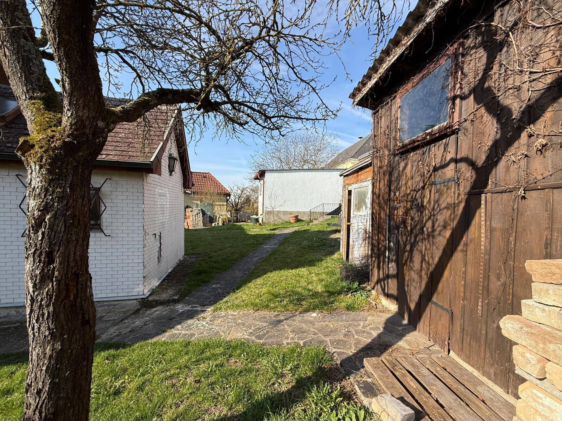 Gartenweg zwischen zwei Nebengebäuden mit Rasenfläche und Bäumen im Hintergrund.