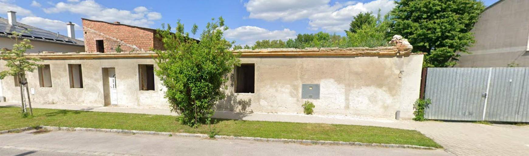 Längere Ansicht der Gebäudefassade mit mehreren Fensteröffnungen und einem Baum im Vorgarten.