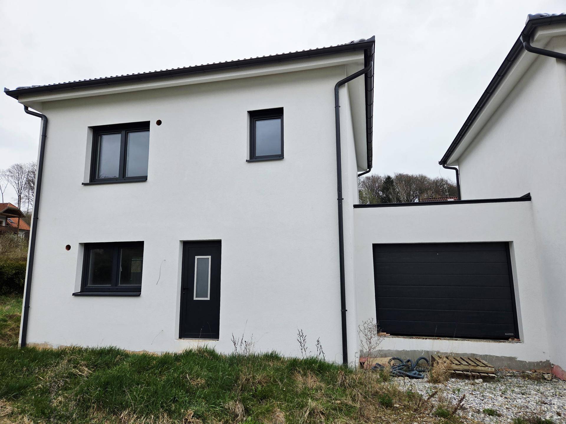 Moderne weiße Hausfassade mit dunklen Fenstern und integrierter Garage unter bewölktem Himmel.