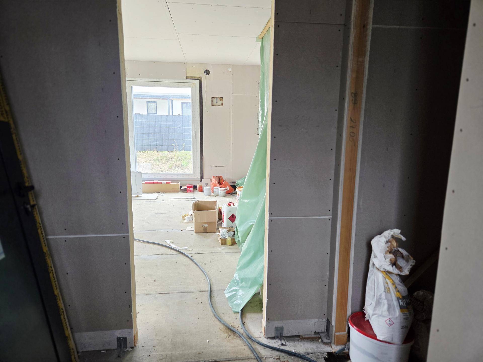 Innenraum im Rohbau mit Trockenbauwänden, großem Fenster und Baumaterialien auf dem Boden.