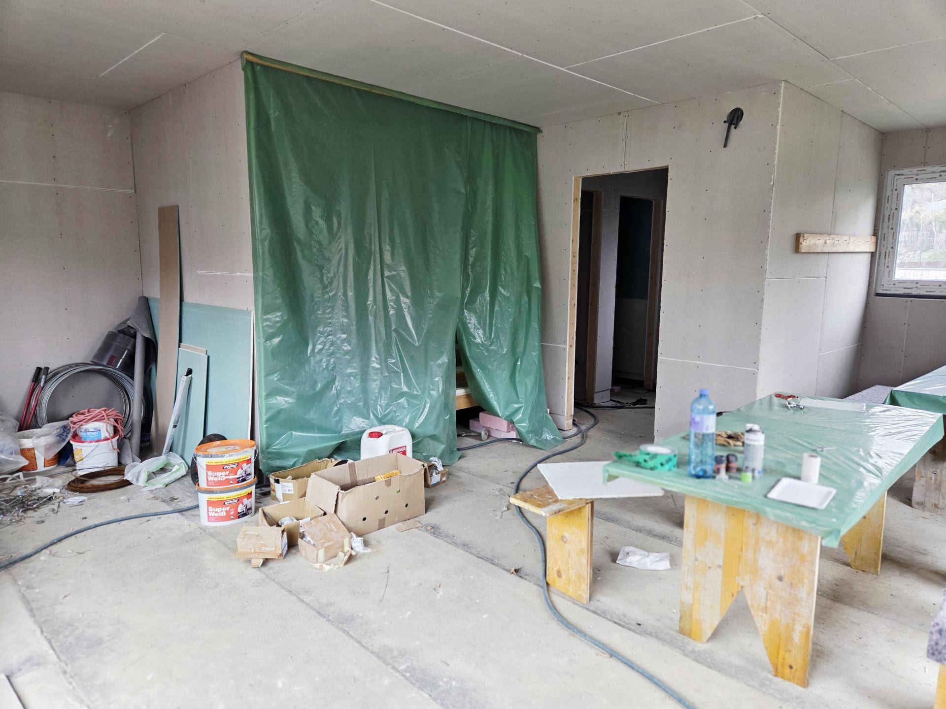 Geräumiger Innenraum im Rohbau mit Trockenbauwänden, grüner Abdeckplane und provisorischen Arbeitstischen.