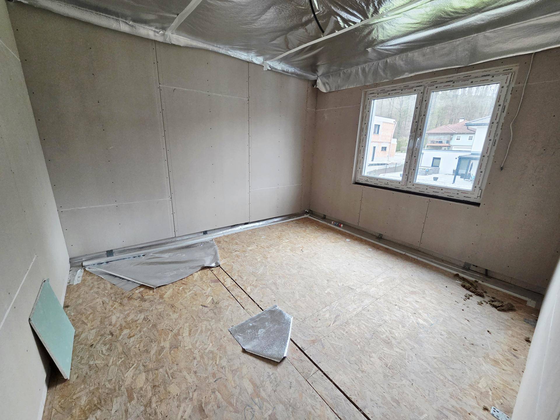 Schlafzimmer im Rohbau mit OSB-Plattenboden, großem Fenster und sichtbarer Deckenisolierung.