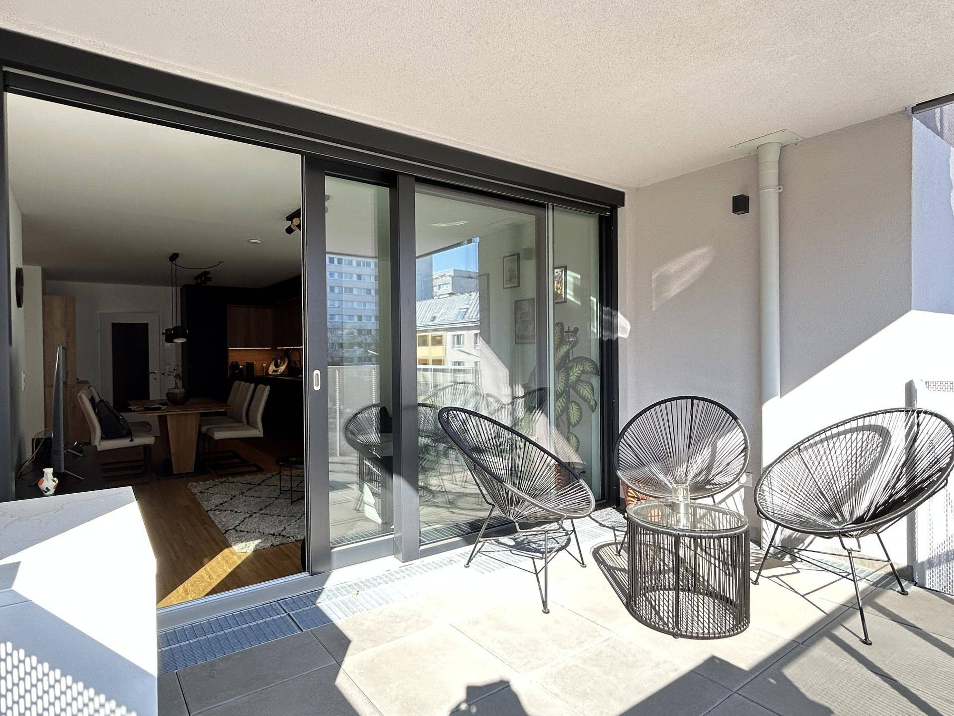Sonniger Balkon mit Fliesenboden, Sitzgelegenheiten und Blick in den Wohnbereich.