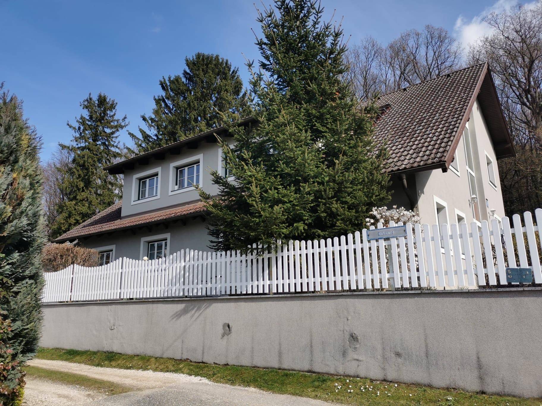 Seitenansicht des Hauses mit weißem Lattenzaun und hohen Nadelbäumen im Hintergrund.
