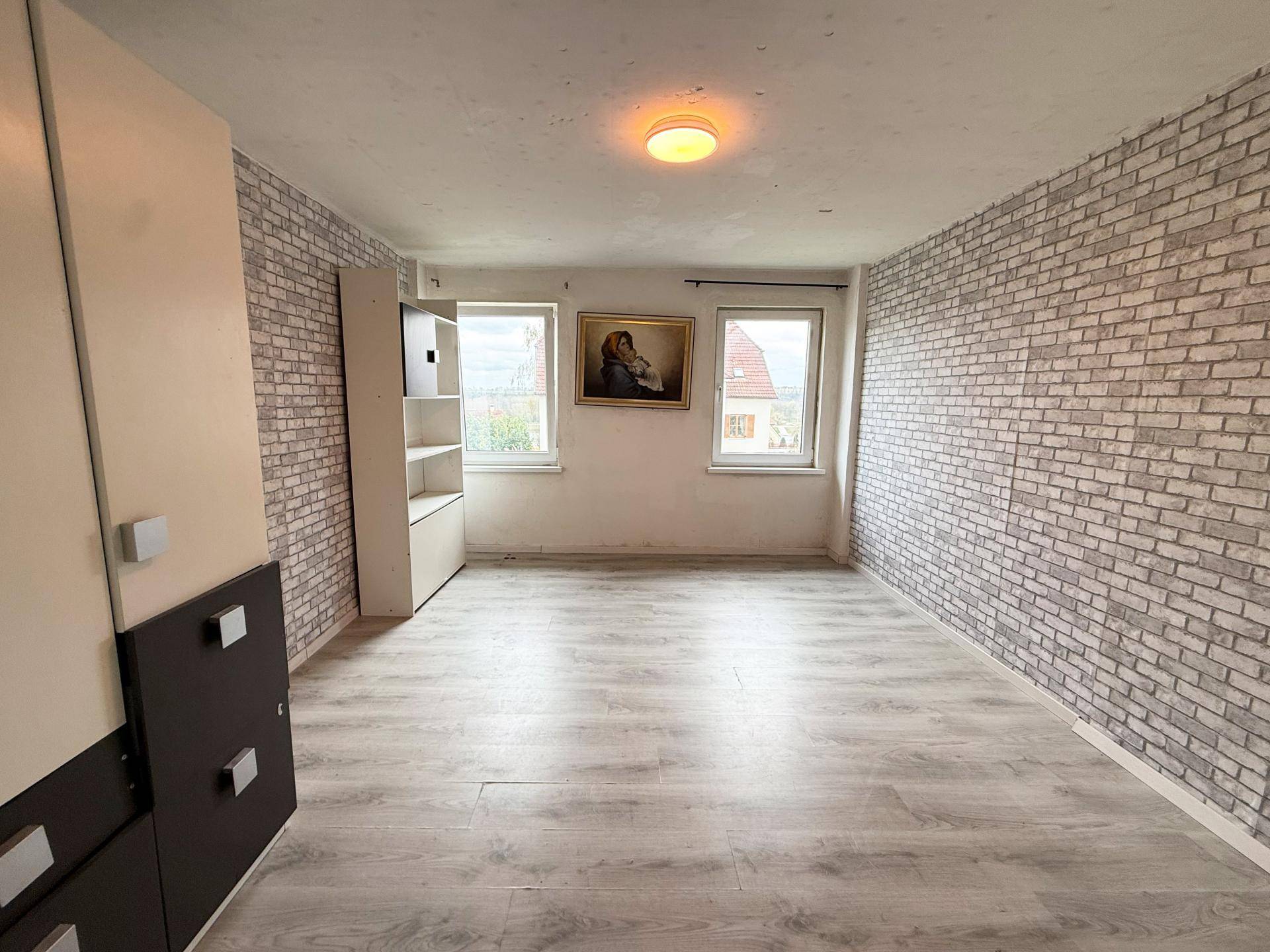 Schlafzimmer mit Laminatboden, zwei Fenstern und einer Wand mit Ziegeloptik-Tapete.