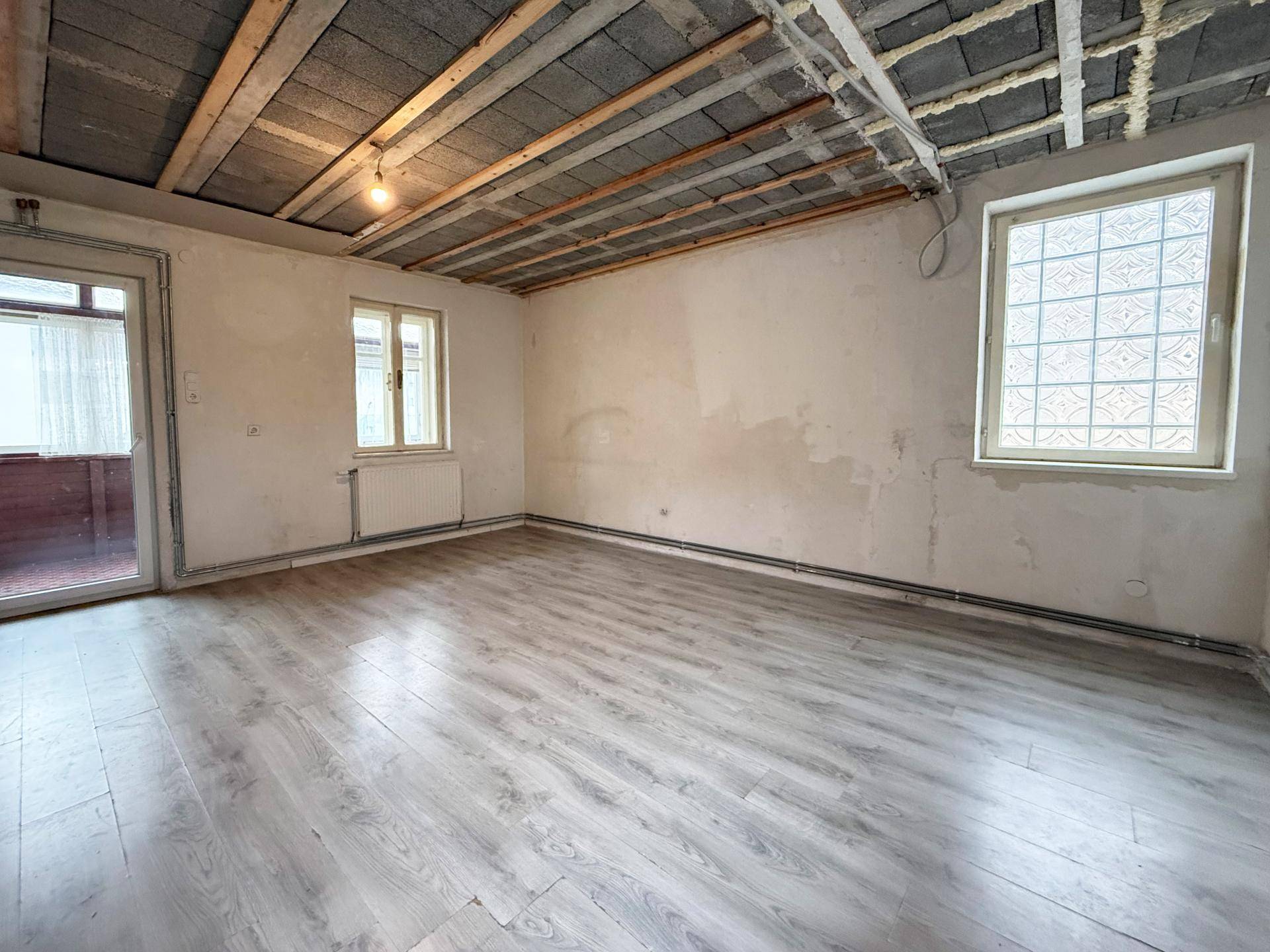 Wohnzimmer im Rohbau mit sichtbarer Deckenkonstruktion und Zugang zur verglasten Loggia.