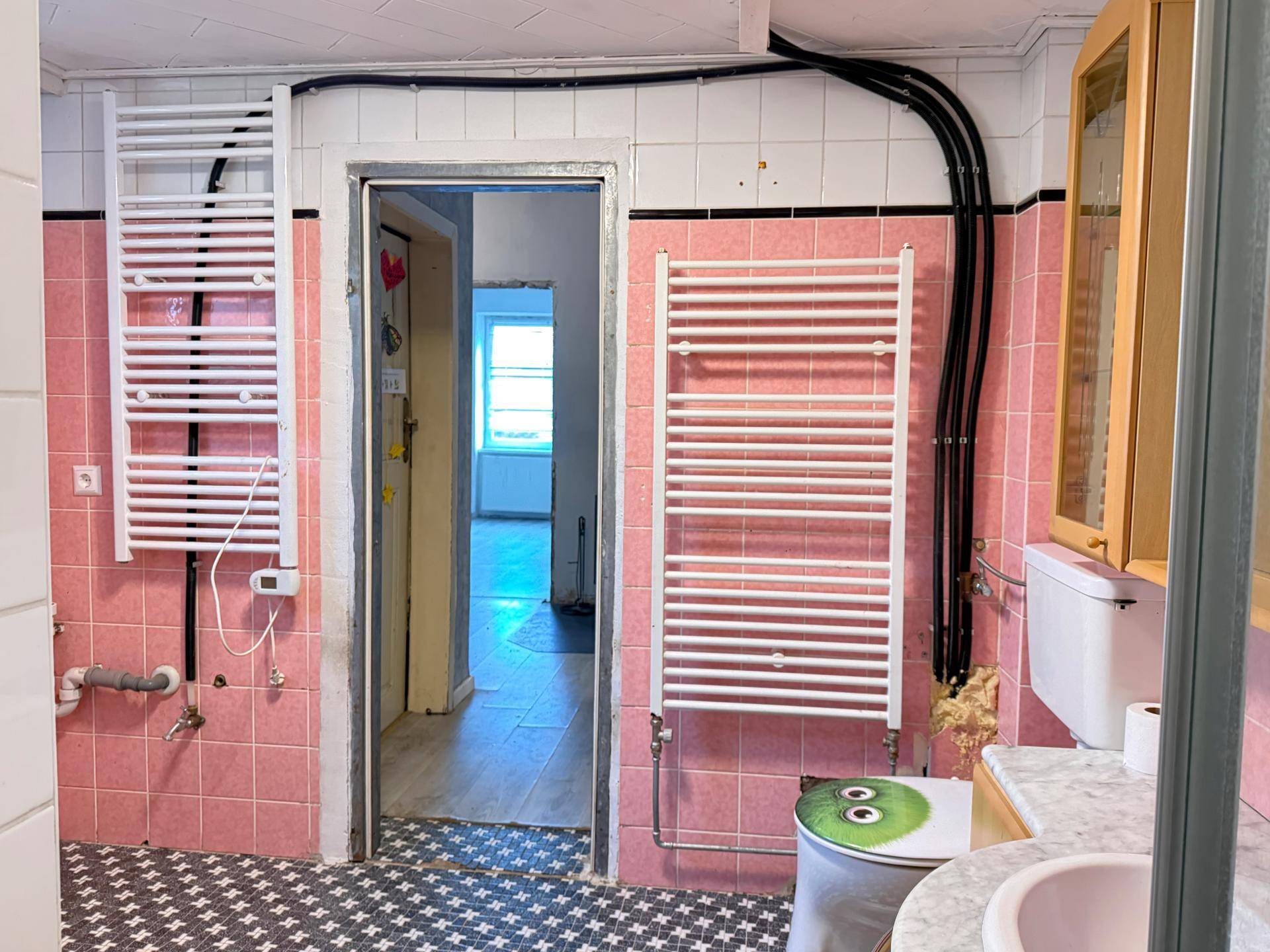 Badezimmer mit rosa Fliesen und zwei Handtuchheizkörpern, bietet Zugang zu einem weiteren Raum.