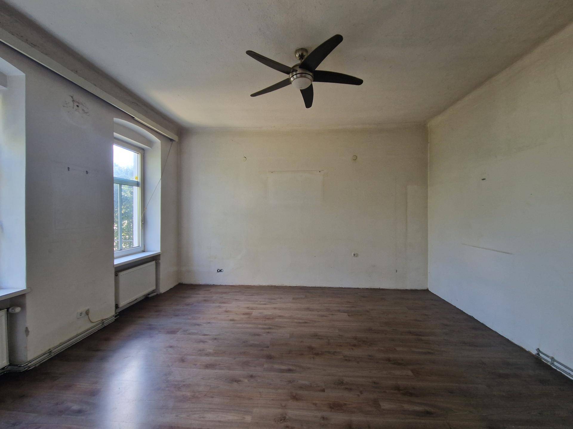 Geräumiges, leeres Zimmer mit Laminatboden, Deckenventilator und zwei Fenstern.