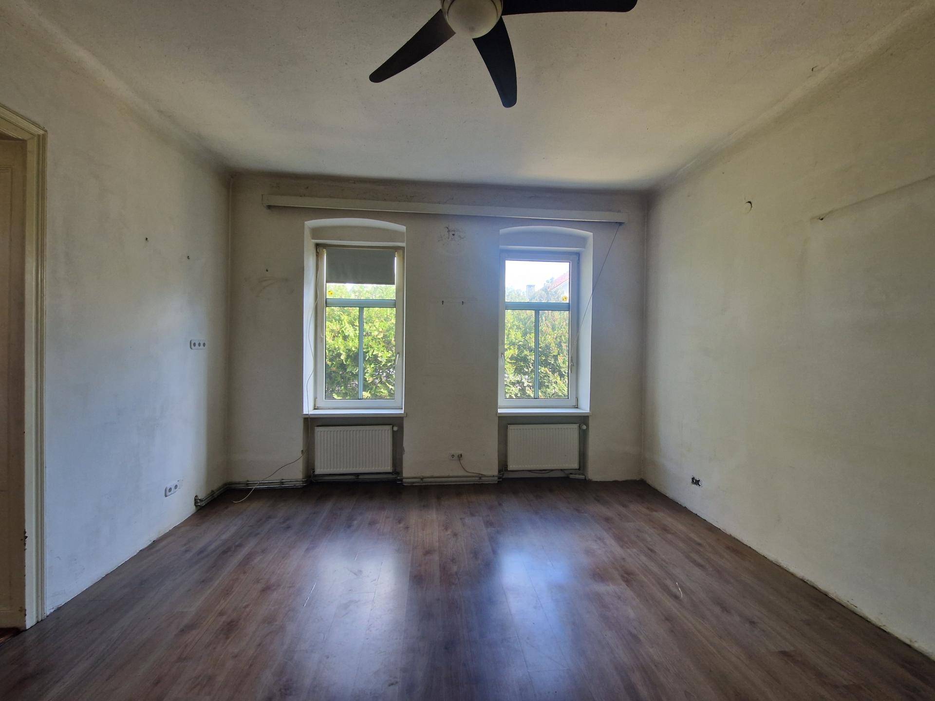 Helles, leeres Zimmer mit Laminatboden, Deckenventilator und Blick ins Grüne durch die Fenster.