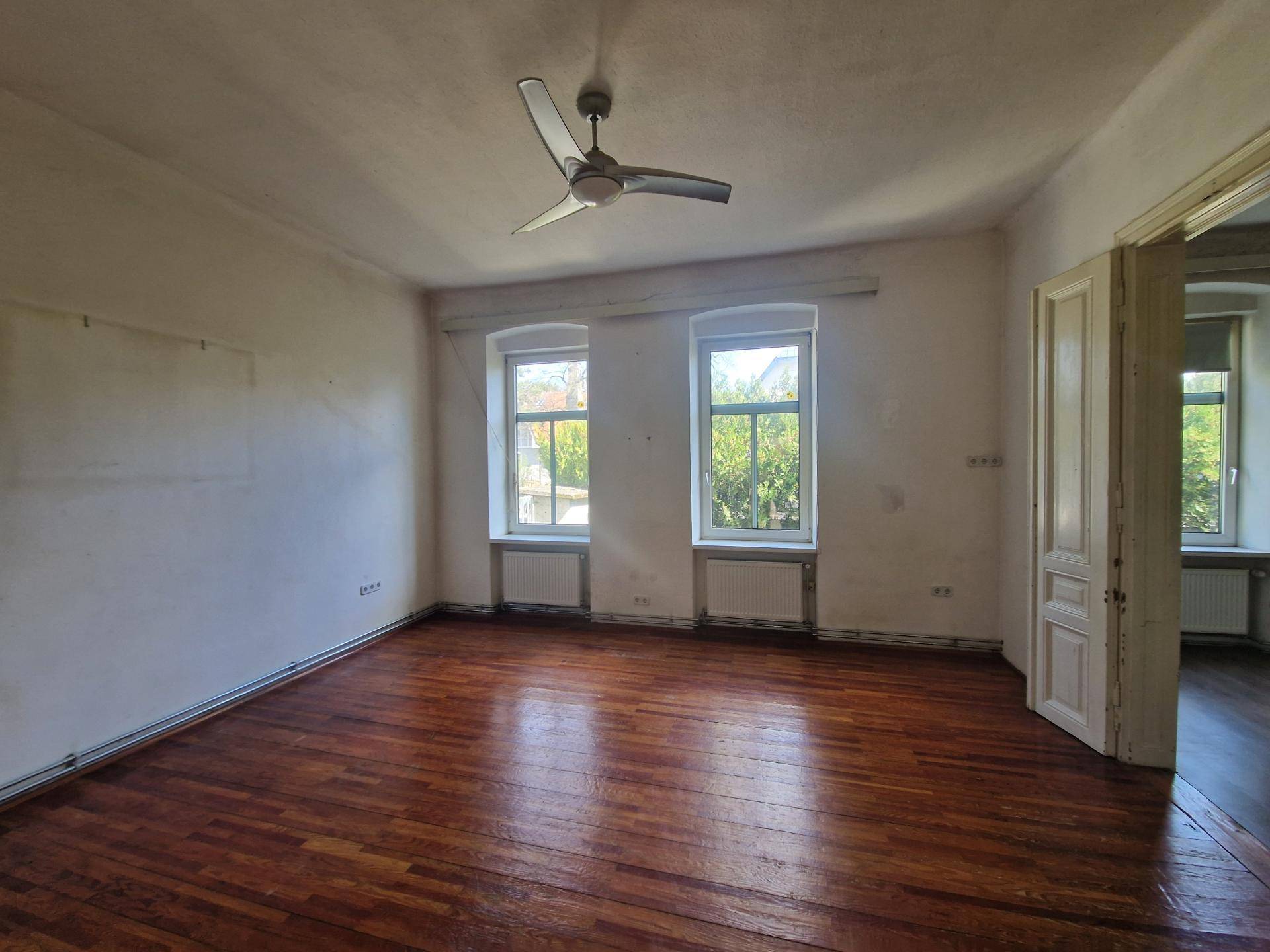 Leeres Zimmer mit dunklem Holzboden, Deckenventilator und Zugang zu einem weiteren Raum.