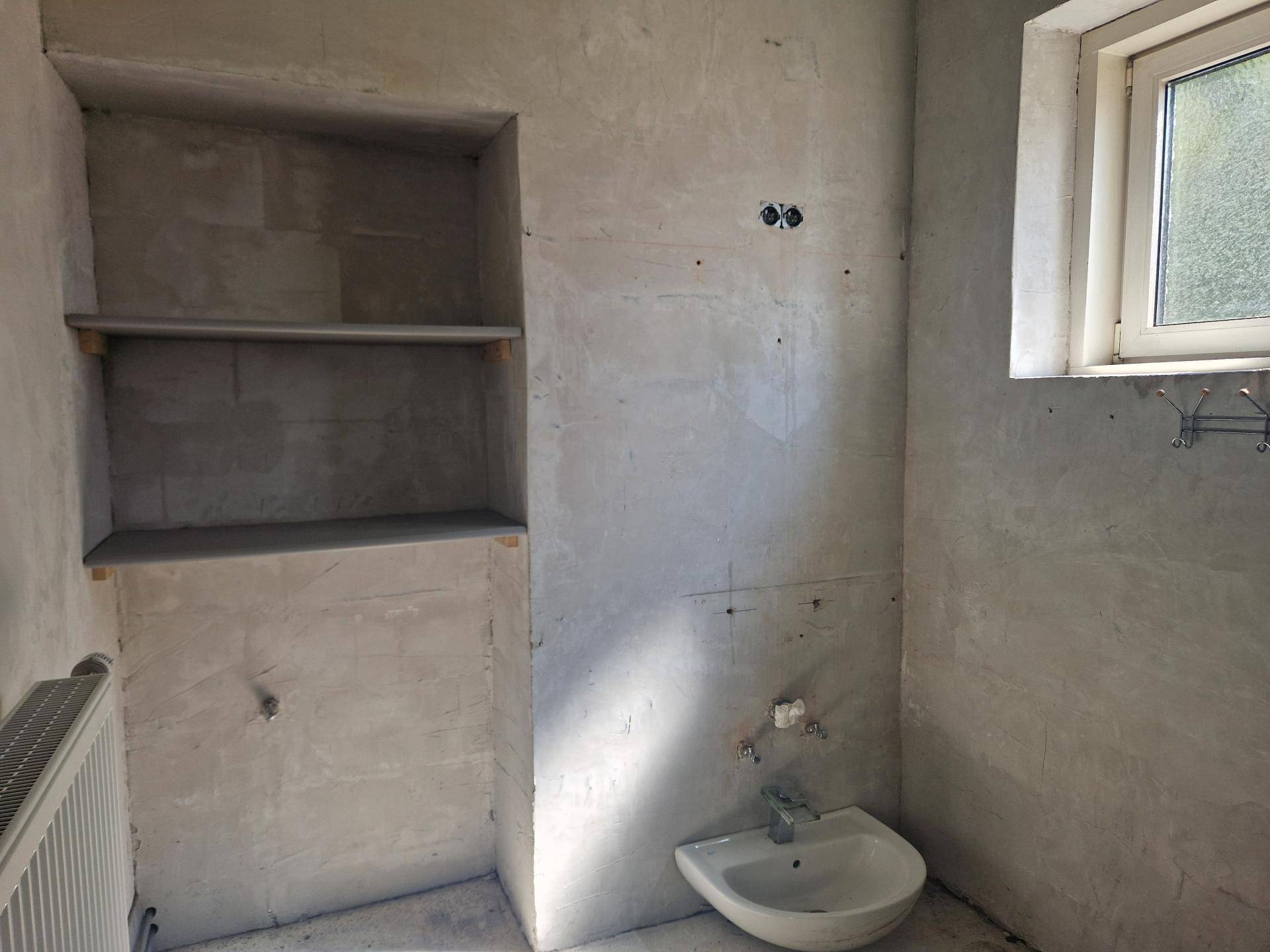Badezimmer im Rohbau mit Waschbecken, Heizkörper und eingebauten Regalen unter einem Fenster.