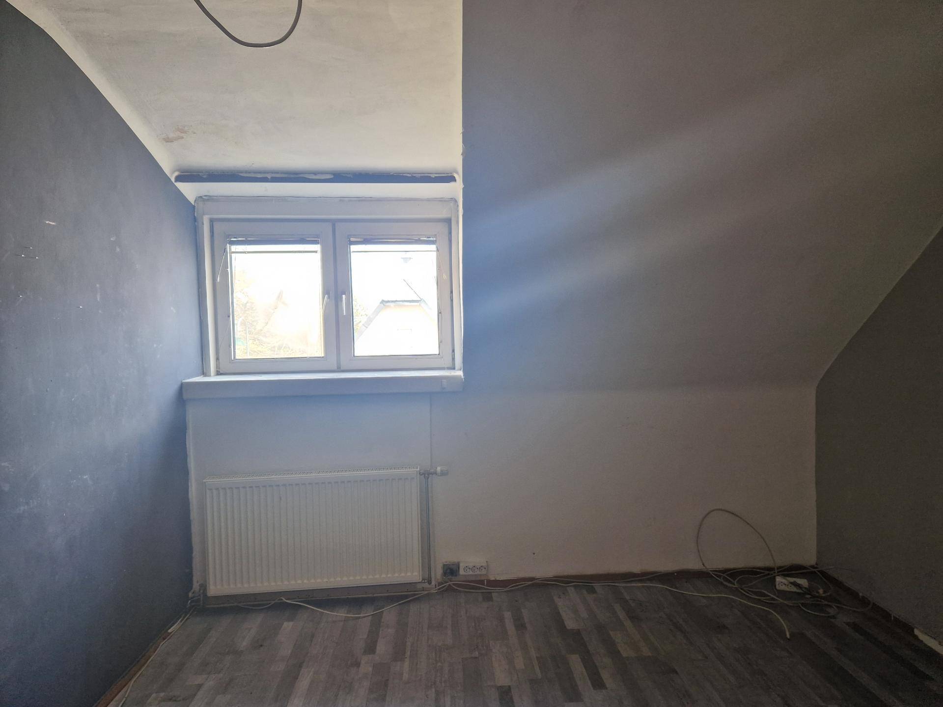 Zimmer im Dachgeschoss mit schrägen Wänden, Fenster und Heizkörper, teilweise gestrichen.