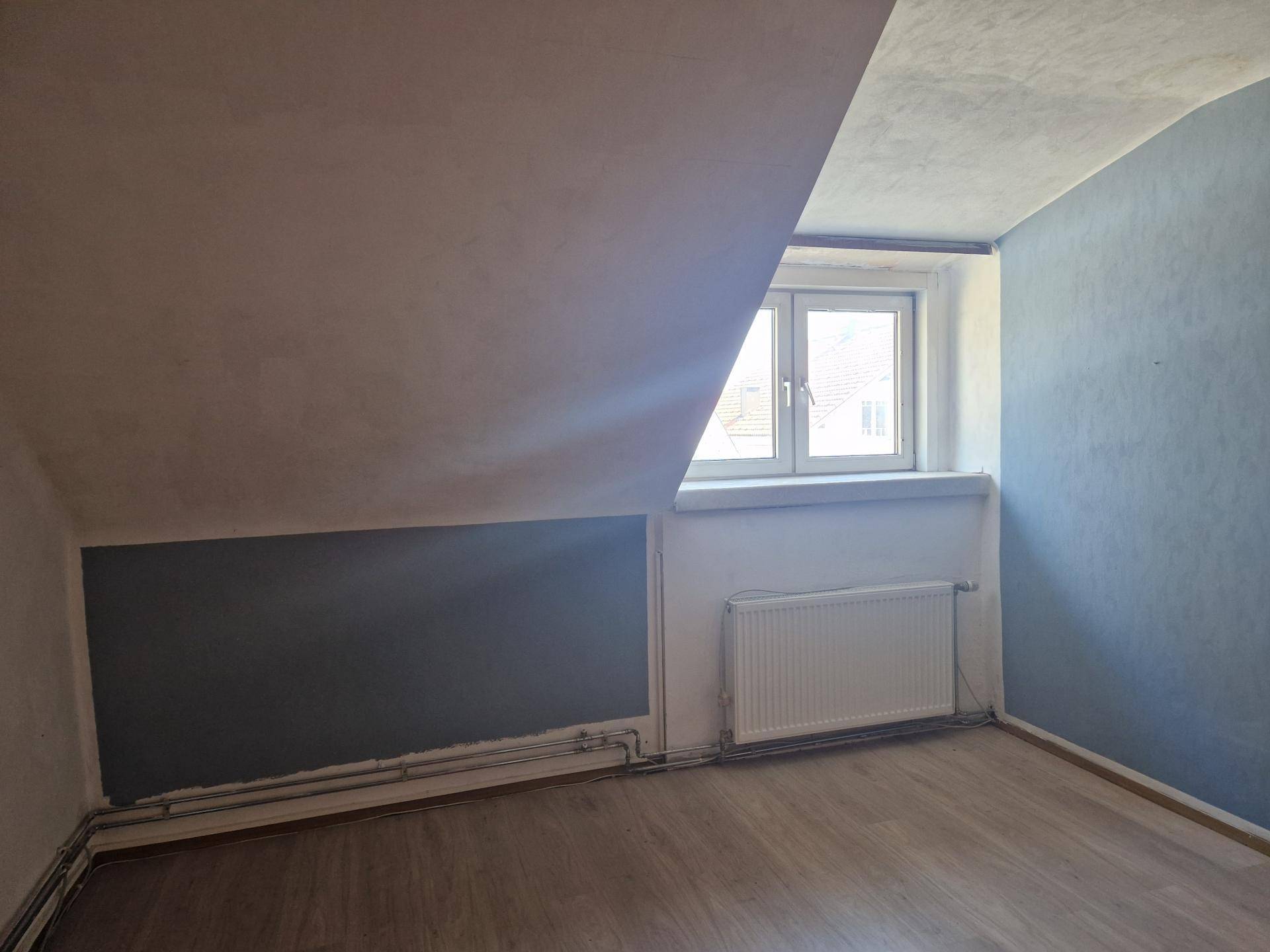Dachgeschosszimmer mit schrägen Wänden, Fenster und Heizkörper, bereit für die Fertigstellung.