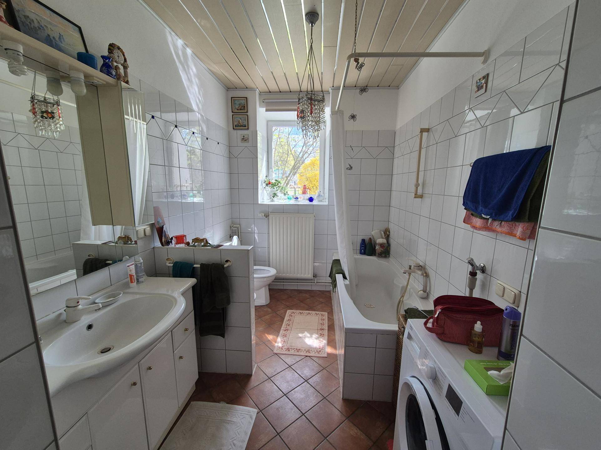 Helles Badezimmer mit Badewanne, Waschbecken, WC und Waschmaschine, gefliesten Wänden und Holzdecke.