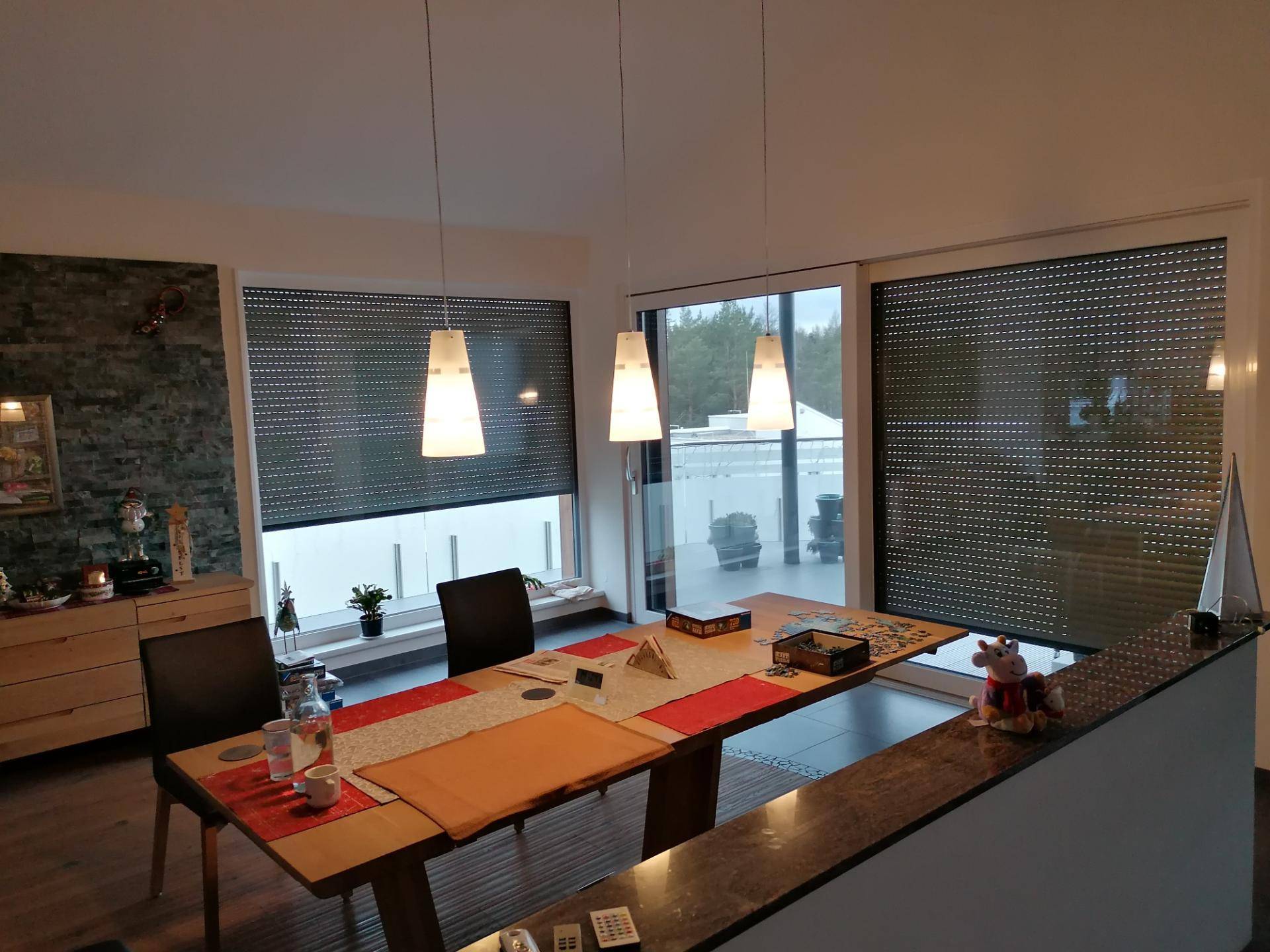Essbereich mit Holztisch, Fliesenboden und Zugang zu einer Terrasse mit Rollläden, hell und modern.