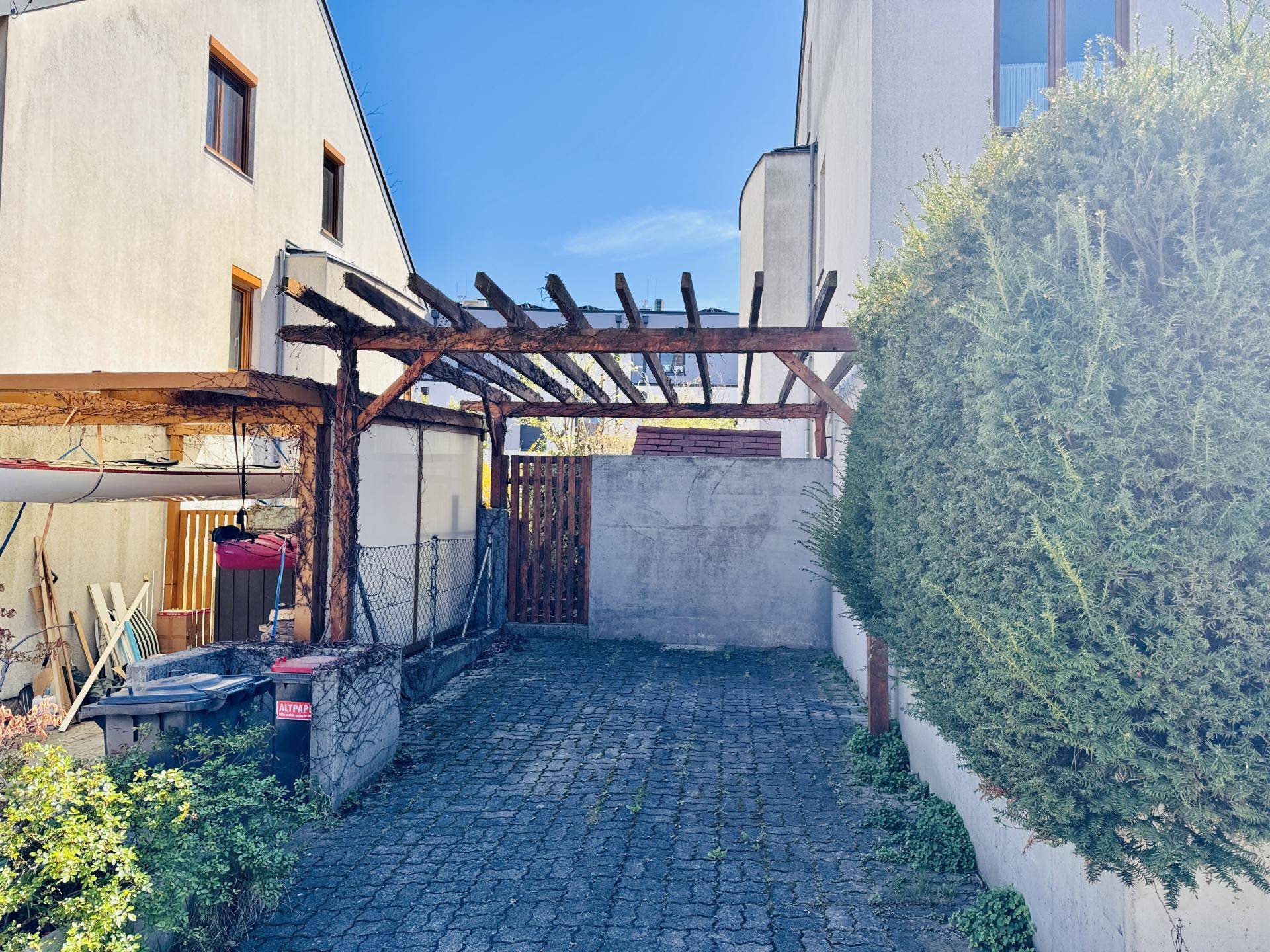 Gepflasterte Einfahrt mit einer Holzpergola und einem Zaun, umgeben von Grünflächen.
