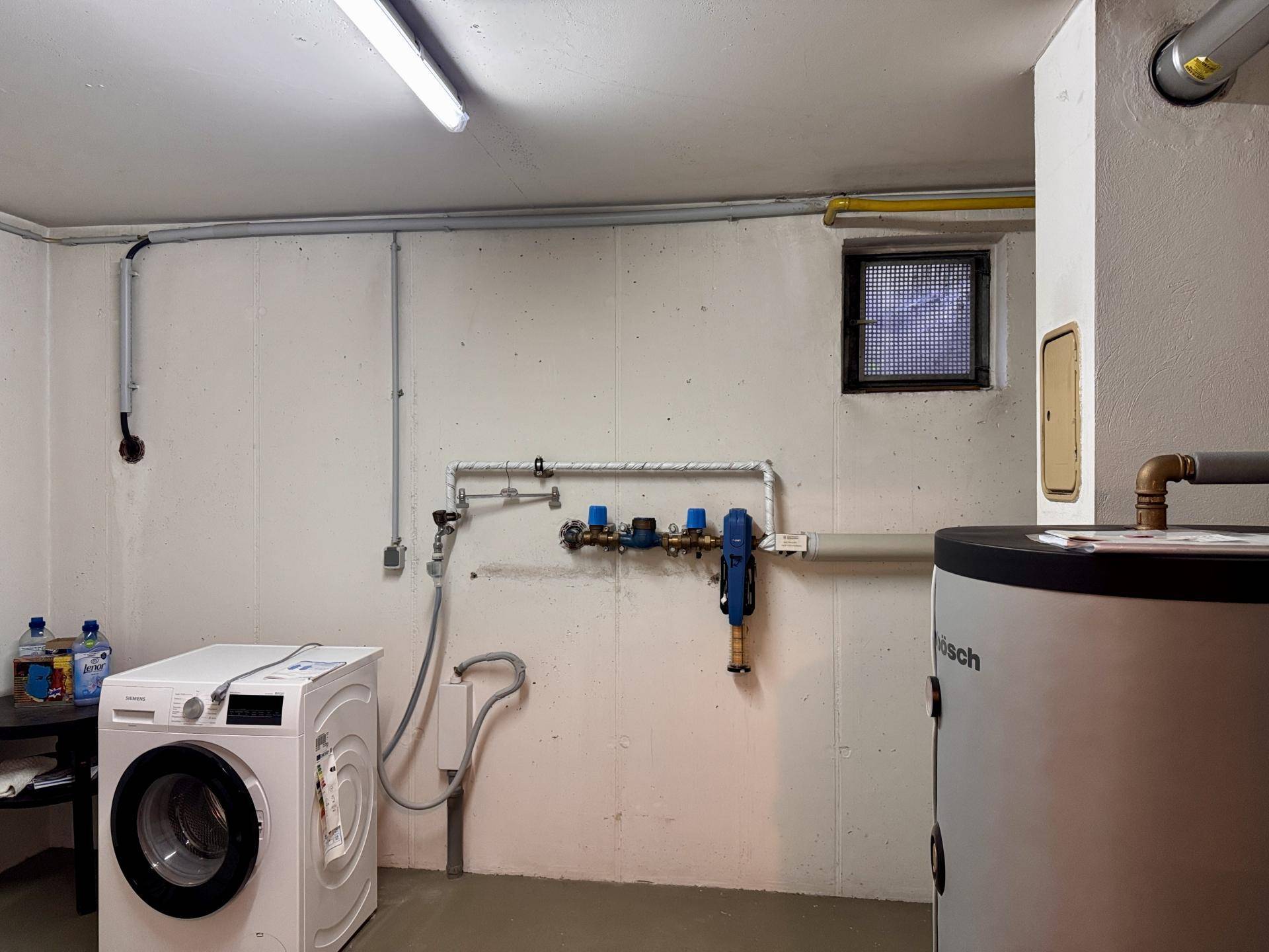 Hauswirtschaftsraum im Keller mit Waschmaschine, Warmwasserbereiter und sichtbaren Installationen.