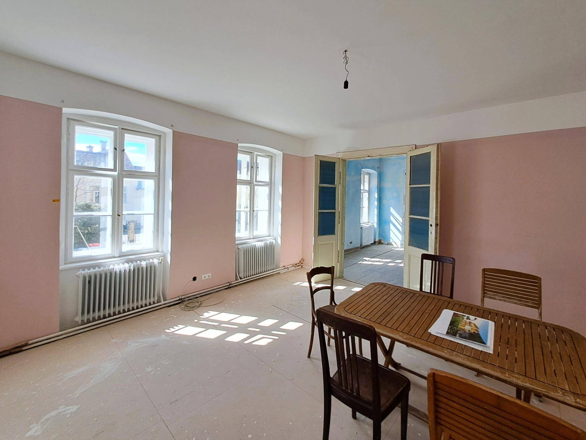 Unmöblierter Raum mit rosa Wänden, hohen Fenstern und einem Holztisch, bereit zur Renovierung.