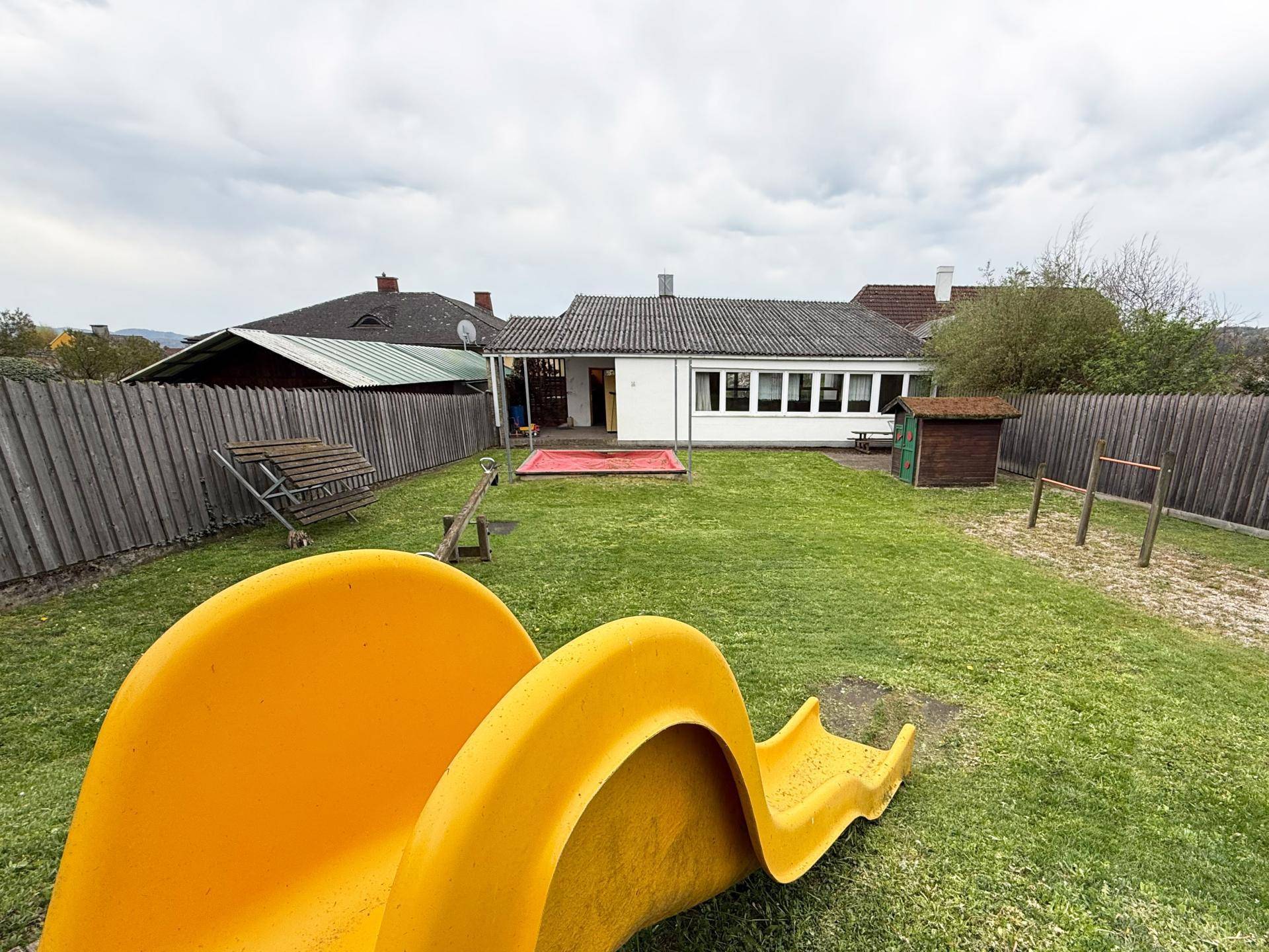 Großer Gartenbereich mit Rasenfläche, Rutsche, Sandkasten und weiteren Spielgeräten für Kinder.