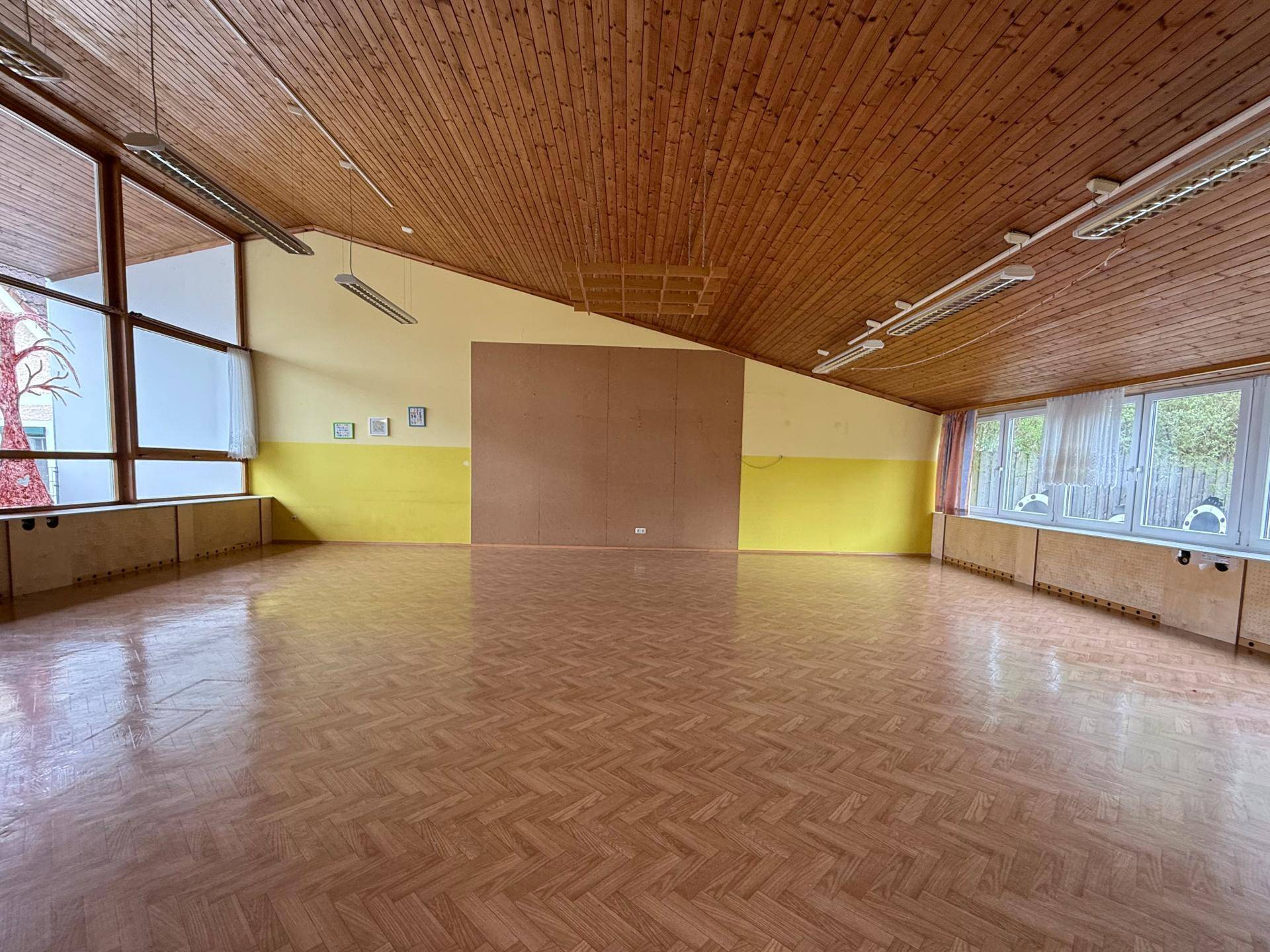 Weitläufiger Raum mit Holzdecke und Parkettboden, bietet viel Platz und Helligkeit durch große Fenster.