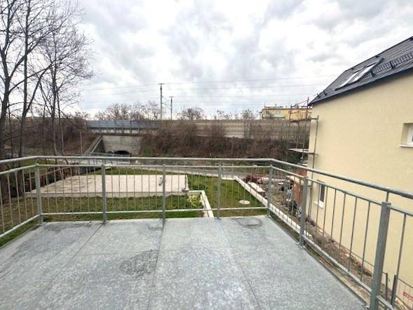 Große Terrasse mit Metallgeländer und Blick auf eine Grünfläche und eine Brücke im Hintergrund.