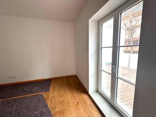 Helles Zimmer mit Holzboden und einer großen Fenstertür, die Zugang zu einem Außenbereich bietet.