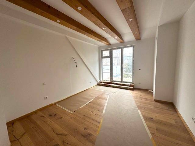 Geräumiges Zimmer mit sichtbaren Holzbalken an der Decke, Holzboden und großen Fenstern.