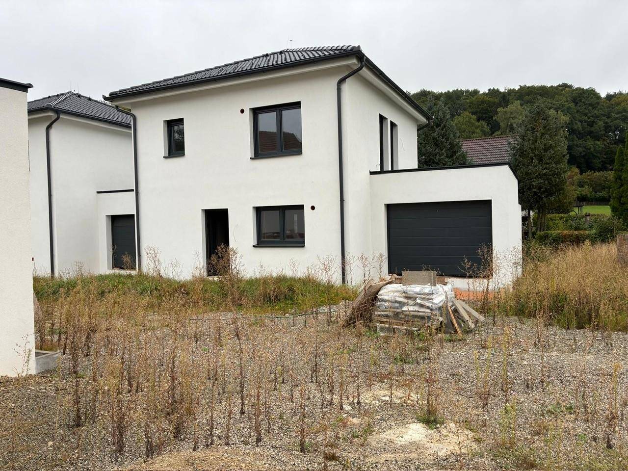 Neubau-Einfamilienhaus mit weißer Fassade, dunklen Fenstern und integrierter Garage, umgeben von unfertigem Gelände.