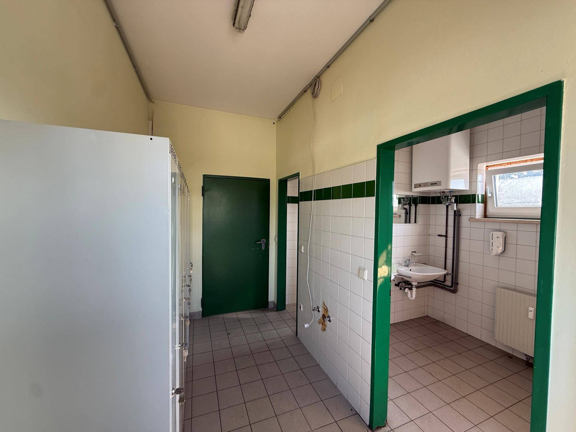 Umkleideraum mit Fliesenboden, Spinden und Zugang zu einem Waschraum mit Fenster für Mitarbeiter.
