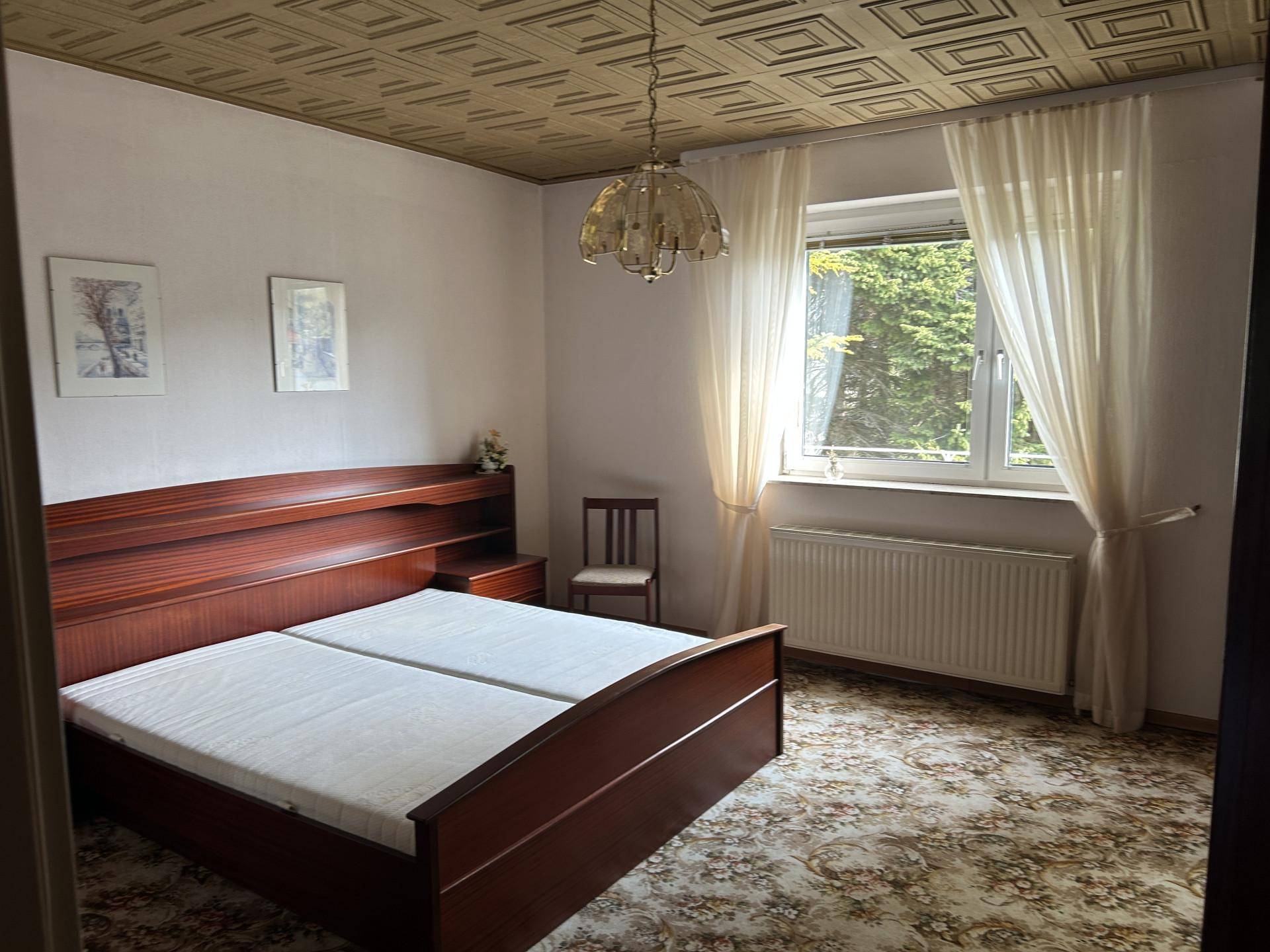 Schlafzimmer mit Doppelbett aus Holz, gemusterter Teppichboden und Fenster zum Garten.