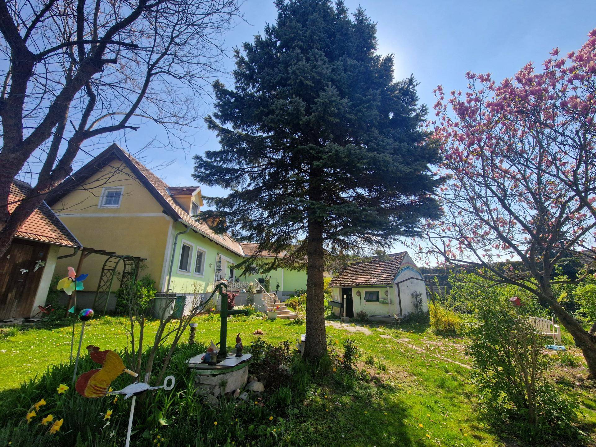 Gepflegter Garten mit hohen Bäumen und verschiedenen Nebengebäuden unter blauem Himmel.