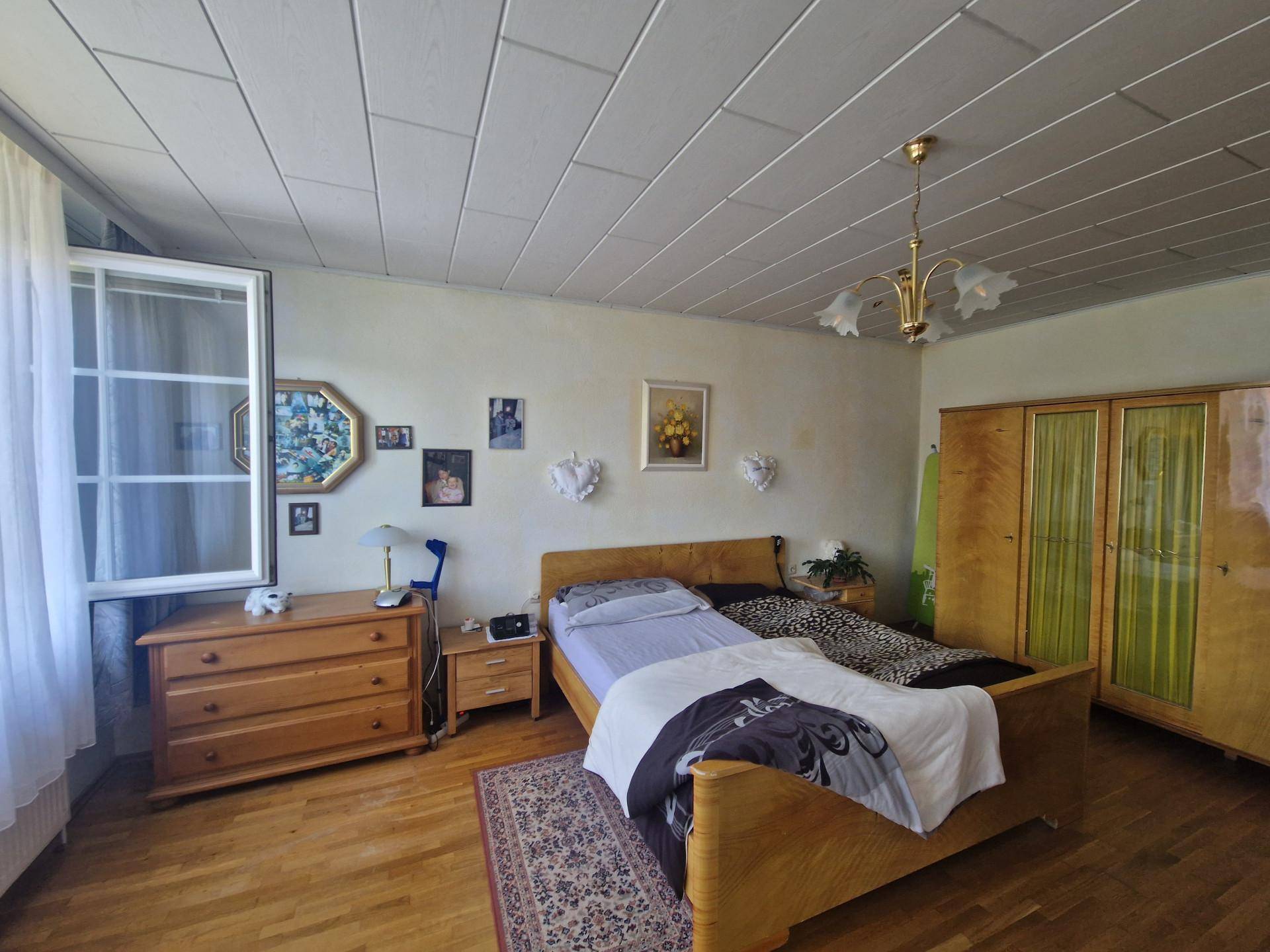 Schlafzimmer mit Doppelbett, Holzschrank und Parkettboden, hell durch großes Fenster.