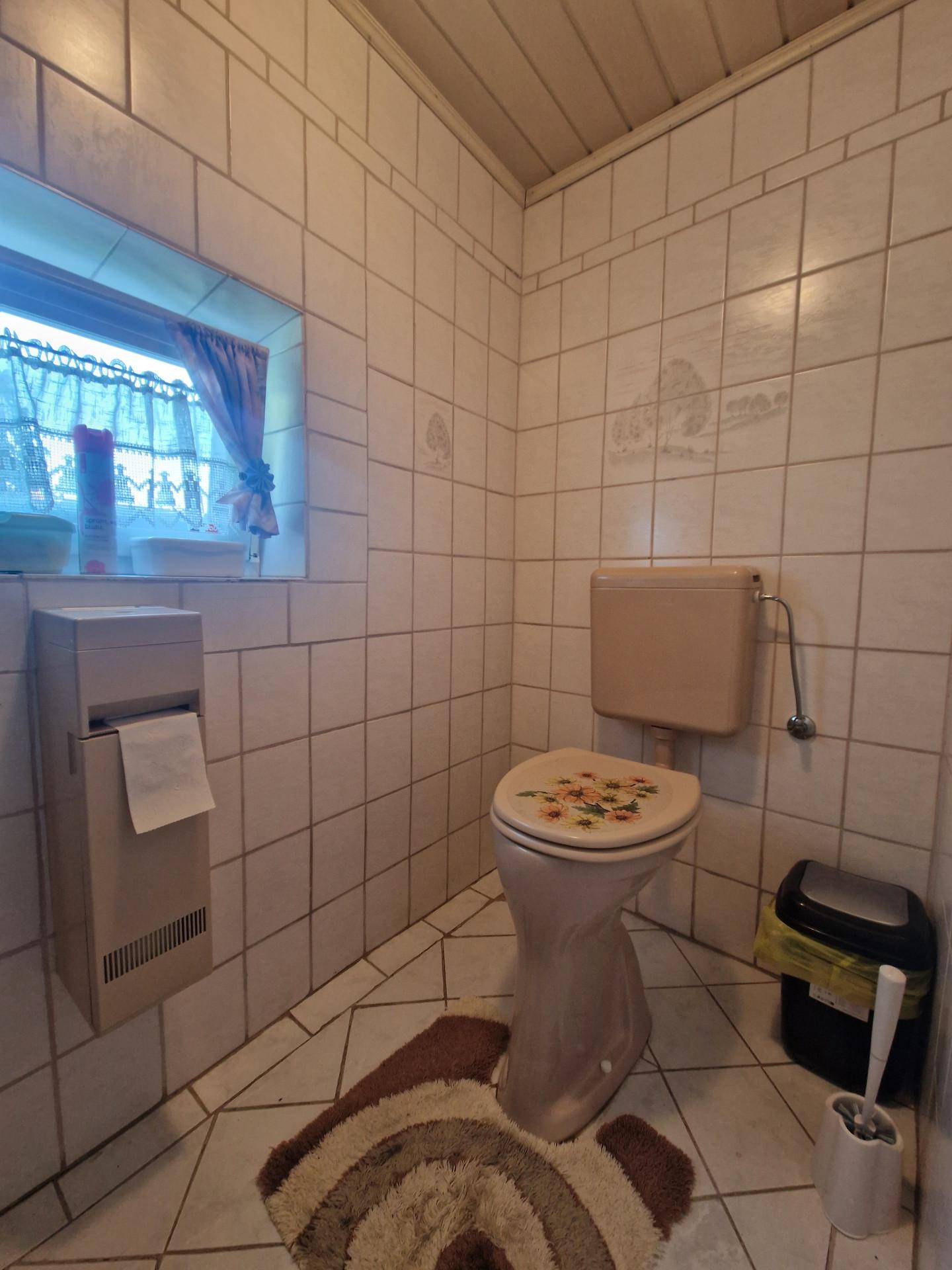 Kleines WC mit beigefarbenen Fliesen, Fenster und traditioneller Toilettenausstattung.