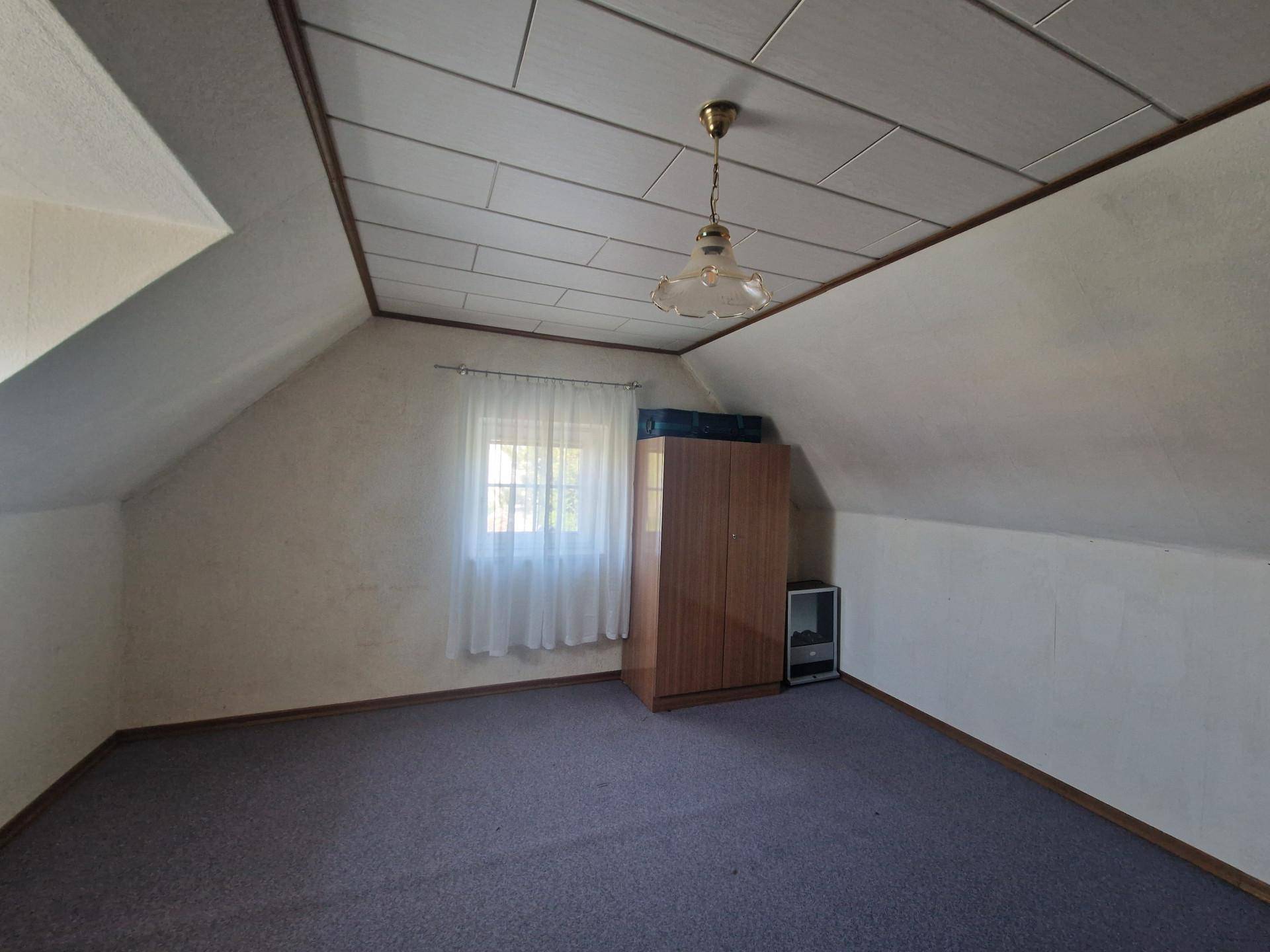 Helles Dachzimmer mit Teppichboden, Dachschräge und einem Fenster mit Blick nach draußen.
