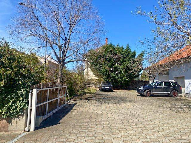 Großer gepflasterter Parkplatz mit Einfahrtstor und Bäumen unter strahlend blauem Himmel.