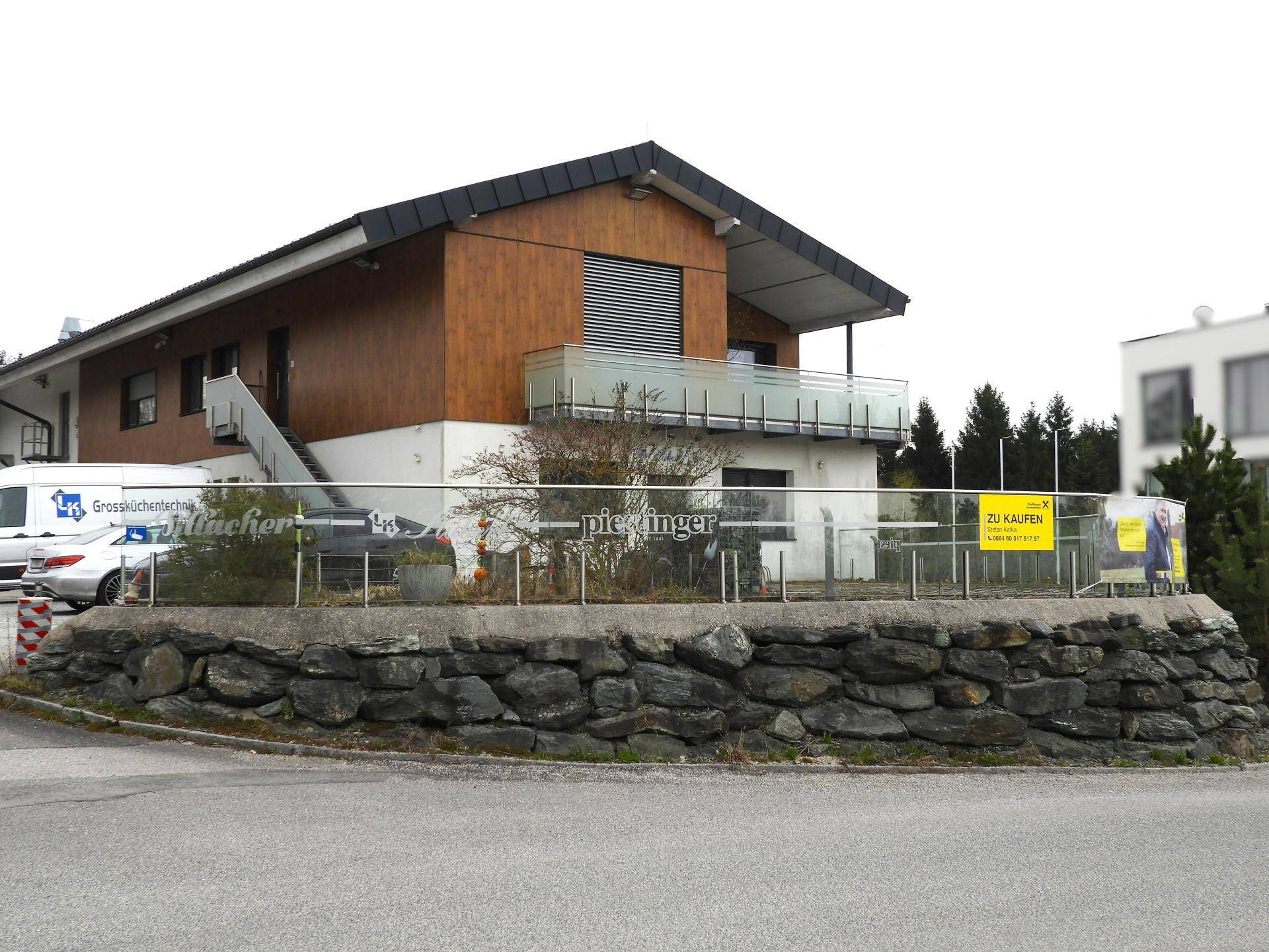 Modernes Gebäude mit Holzfassade, Balkon und Steinfundament, umgeben von Natur und Parkplätzen.