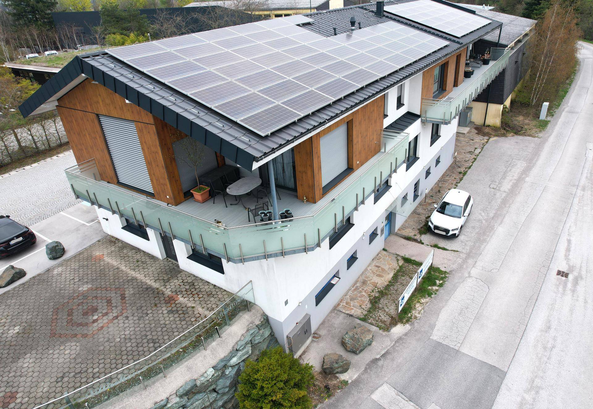 Luftaufnahme eines modernen Gebäudes mit Photovoltaikanlage und möblierter Terrasse, umgeben von Natur.