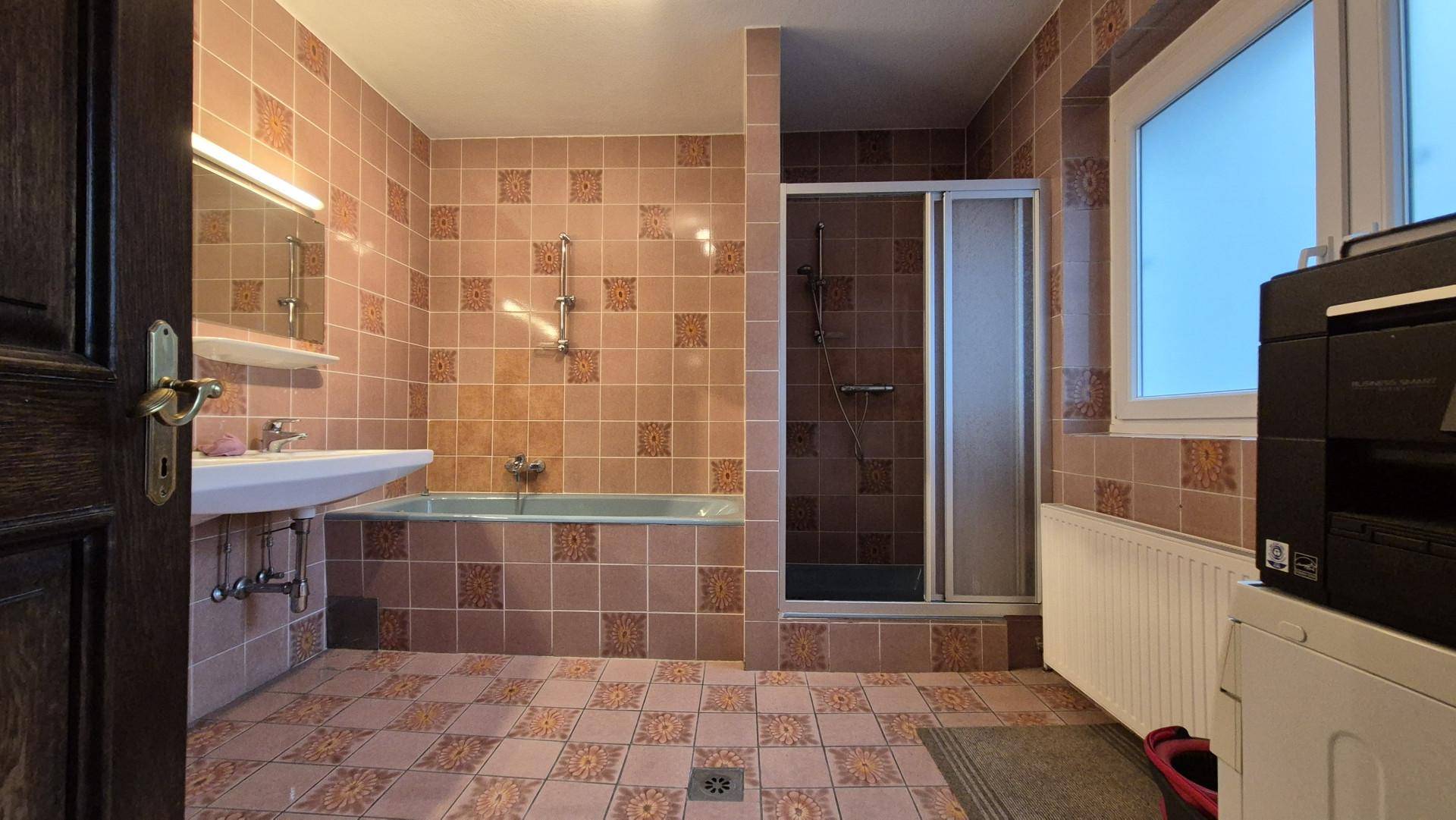 Badezimmer mit Badewanne, Dusche, Waschbecken und gemusterten Fliesen im 80er-Jahre-Stil.