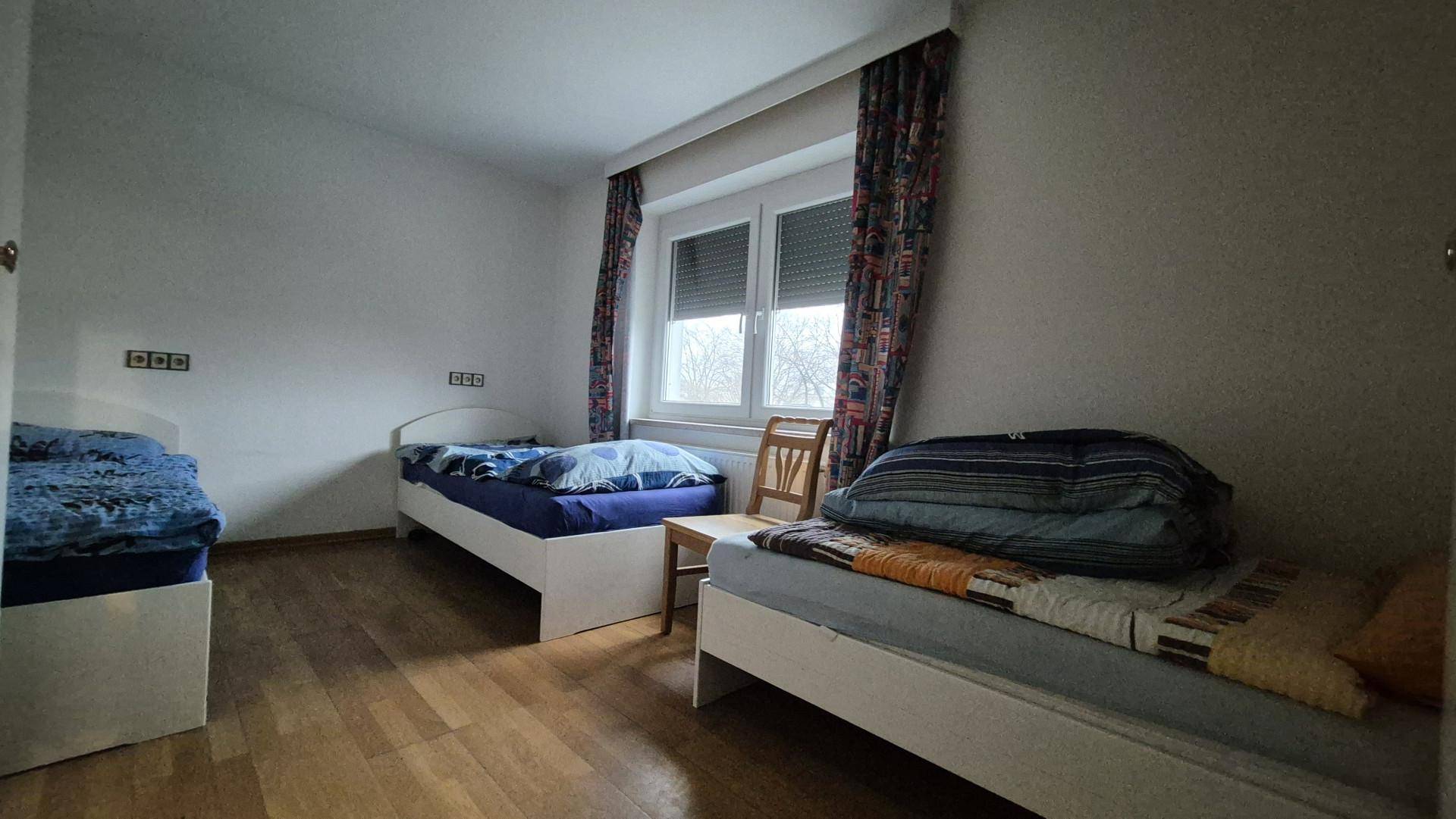 Schlafzimmer mit drei Einzelbetten und Holzboden, gut beleuchtet durch ein großes Fenster.